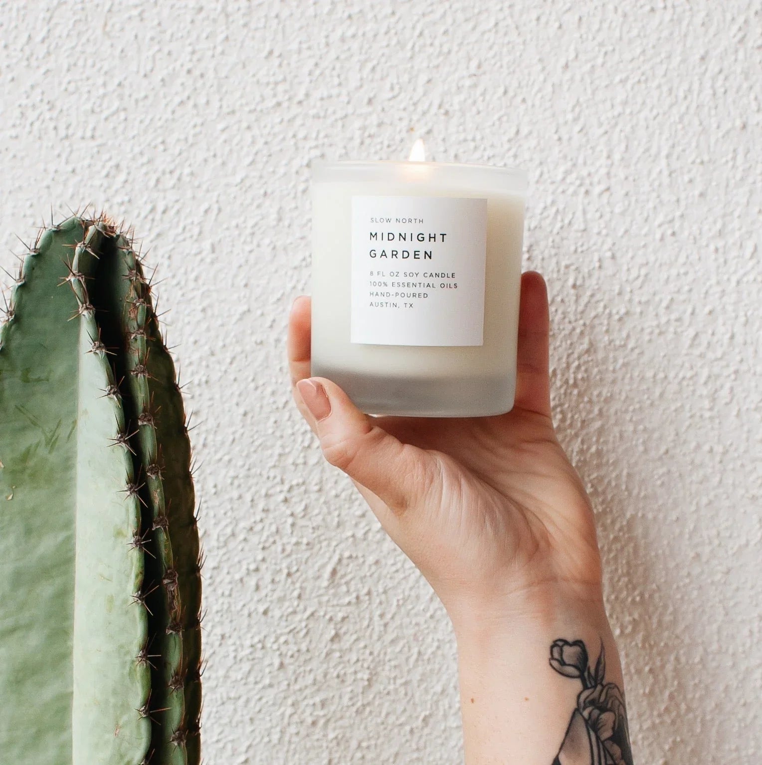 Hand holding lit Midnight Garden soy candle with tattooed wrist near green cactus on textured wall