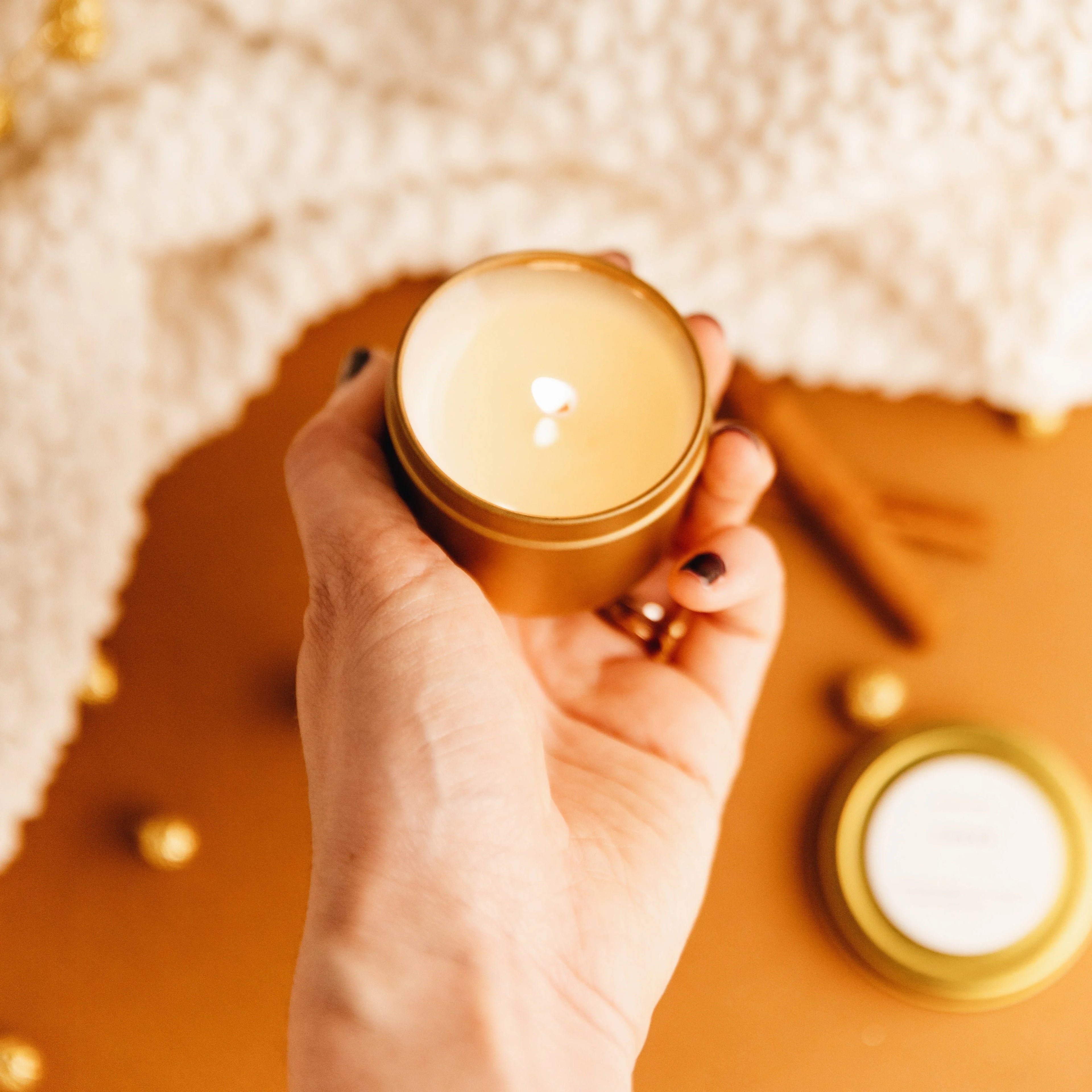 Hand holding a lit mini 2 oz scented candle in a gold tin with warm, cozy background