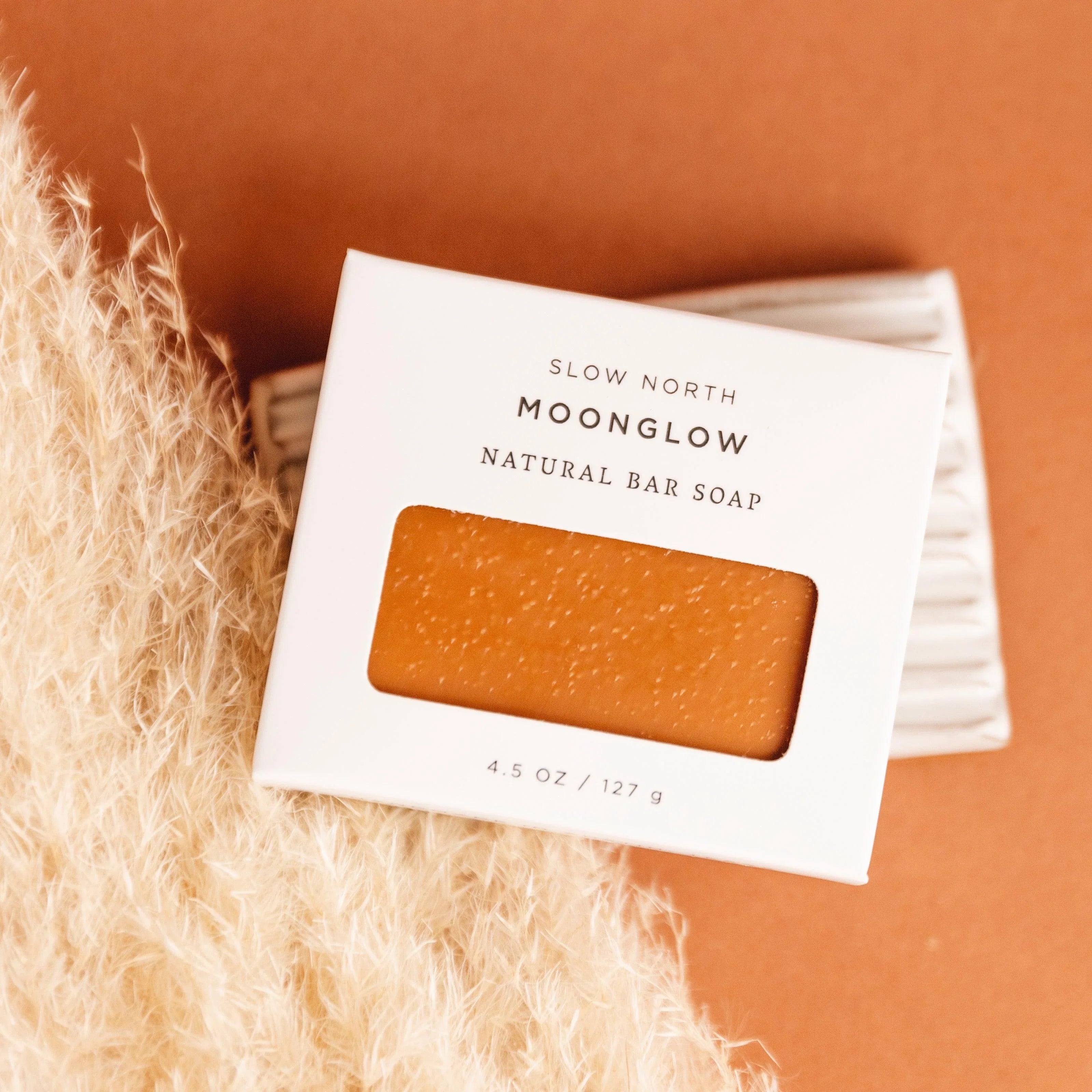Slow North Moonglow natural bar soap in white box with transparent window on orange background and pampas grass