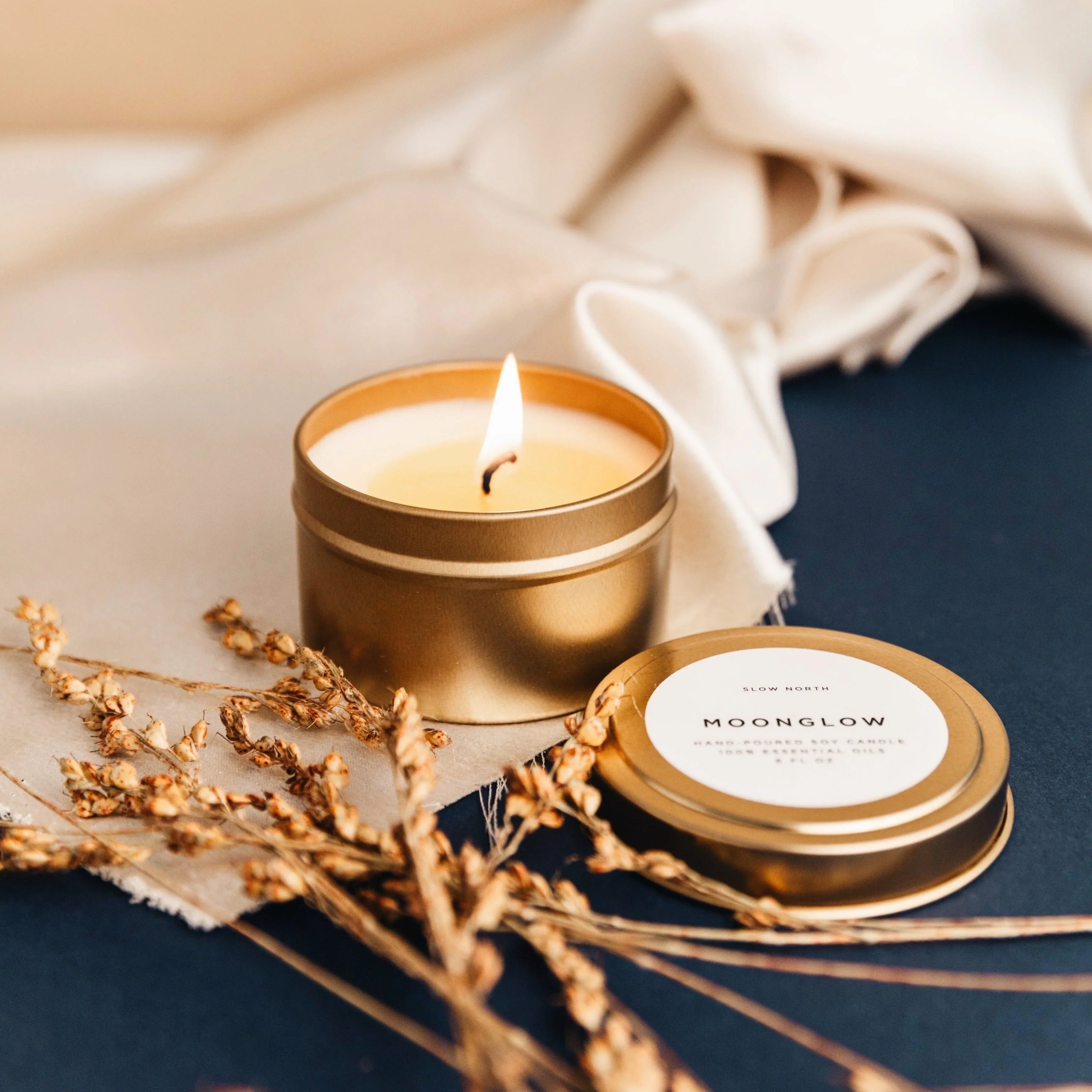 Moonglow travel tin soy candle burning beside lid and dried flowers on fabric and dark surface