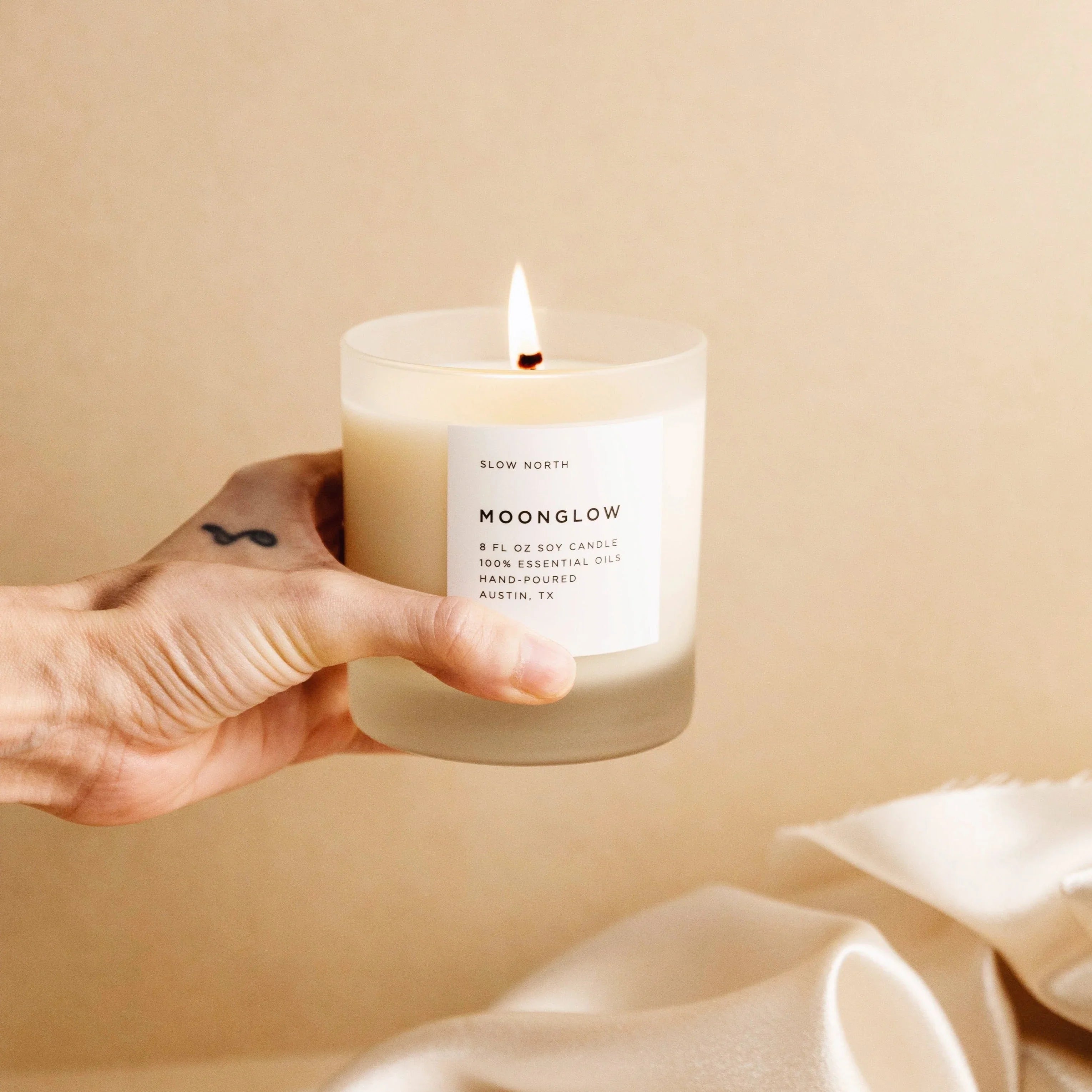 Hand holding lit Moonglow soy candle in frosted glass with beige background and fabric