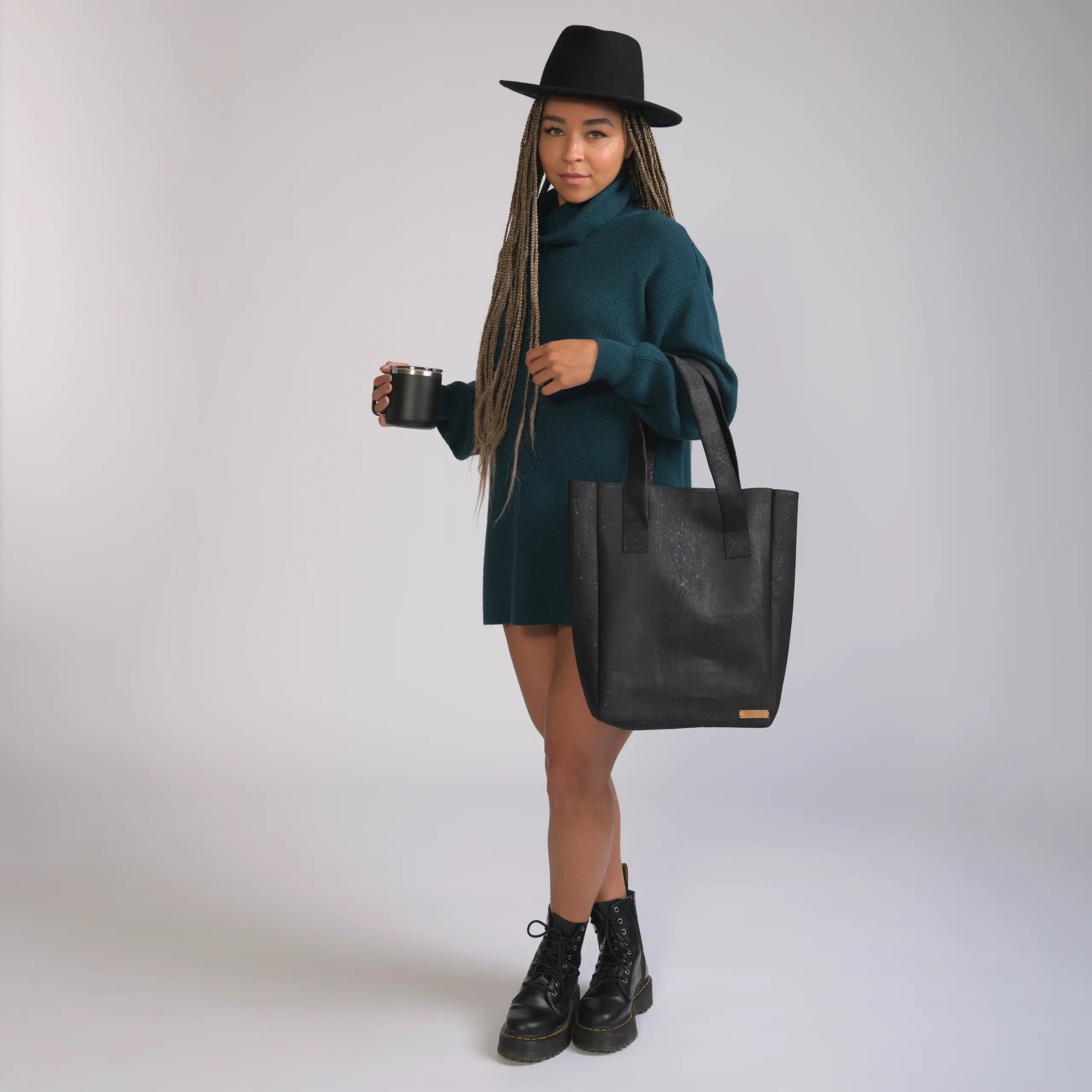 Woman in teal sweater dress, black hat, and boots holding a black tote bag from Qu!rk Boutique