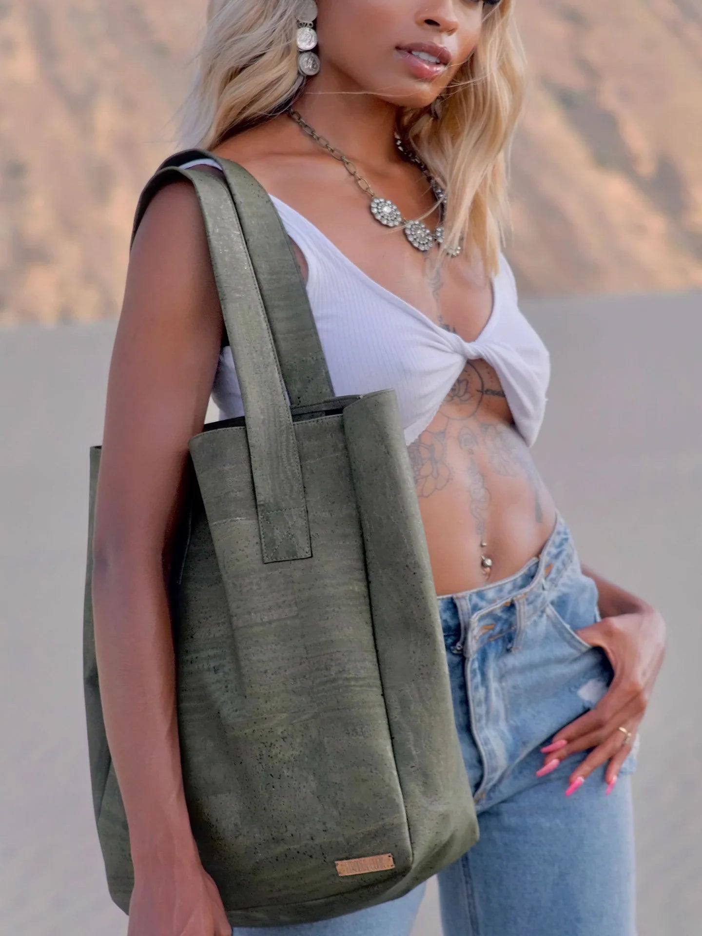 Woman modeling a large olive green tote bag, wearing a white top and jeans, with desert backdrop