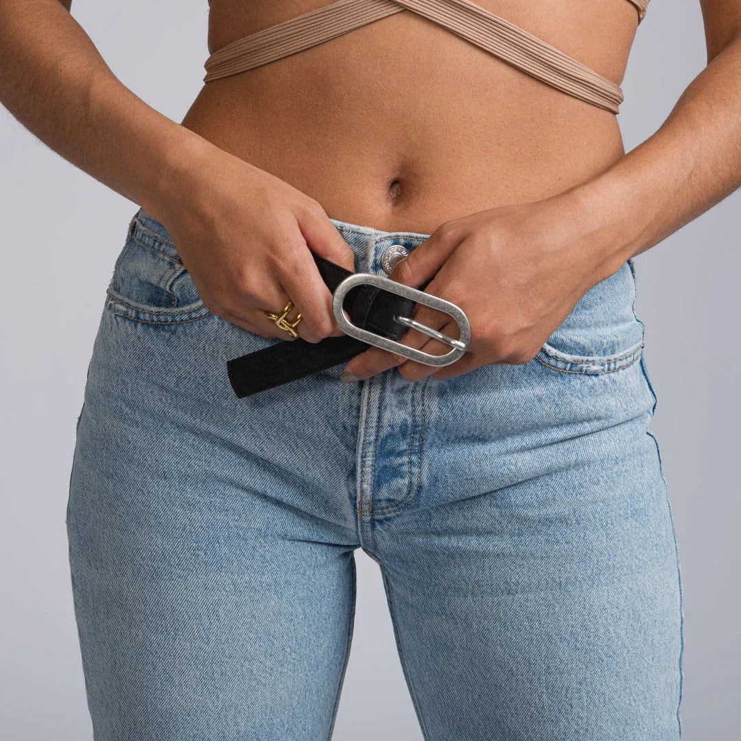 Woman wearing high-rise blue jeans and taupe crop top adjusts large silver belt buckle