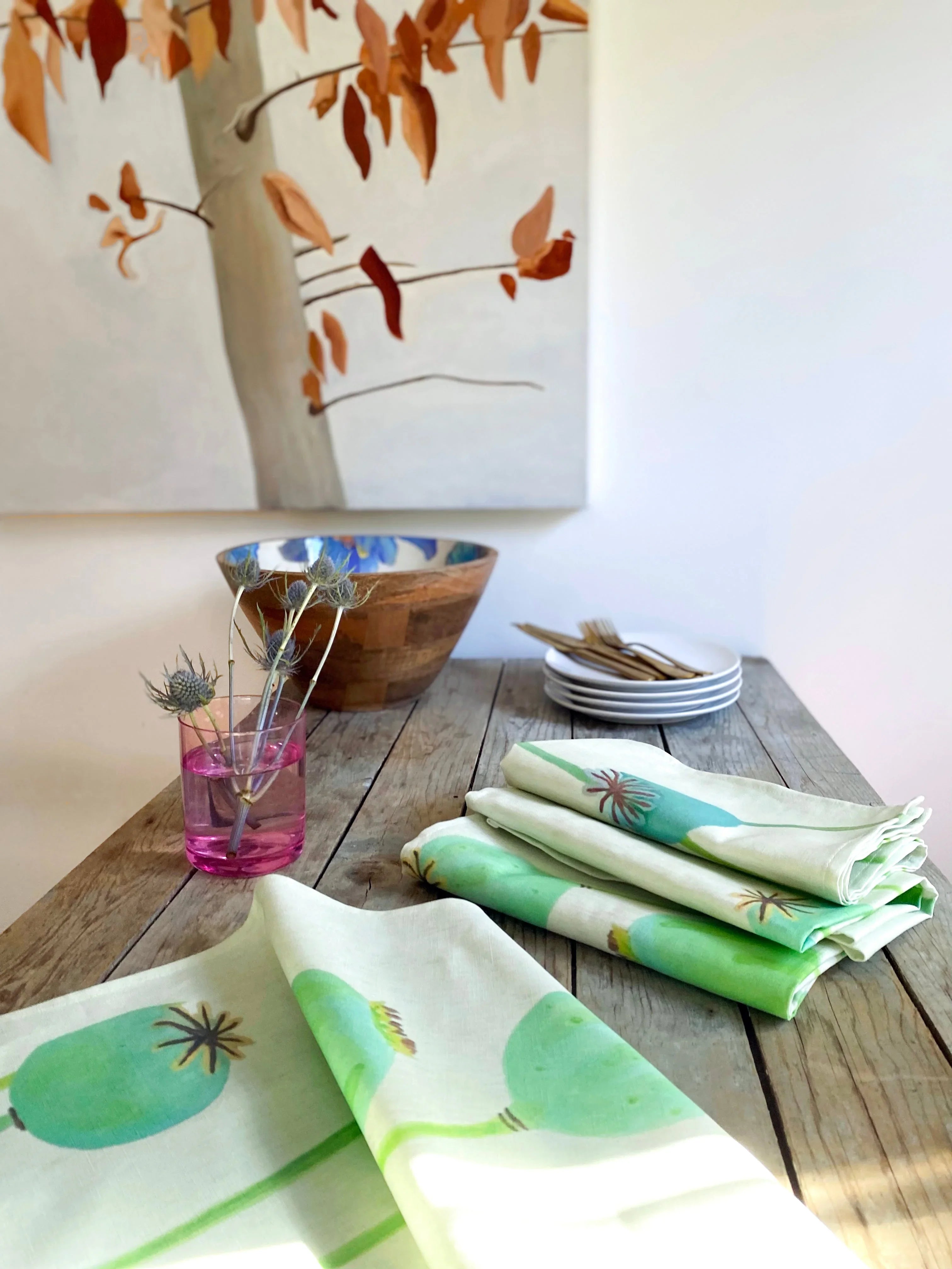 Ecru napkins with green poppy pods on rustic wooden table with pink glass vase, wooden bowl, and stacked plates