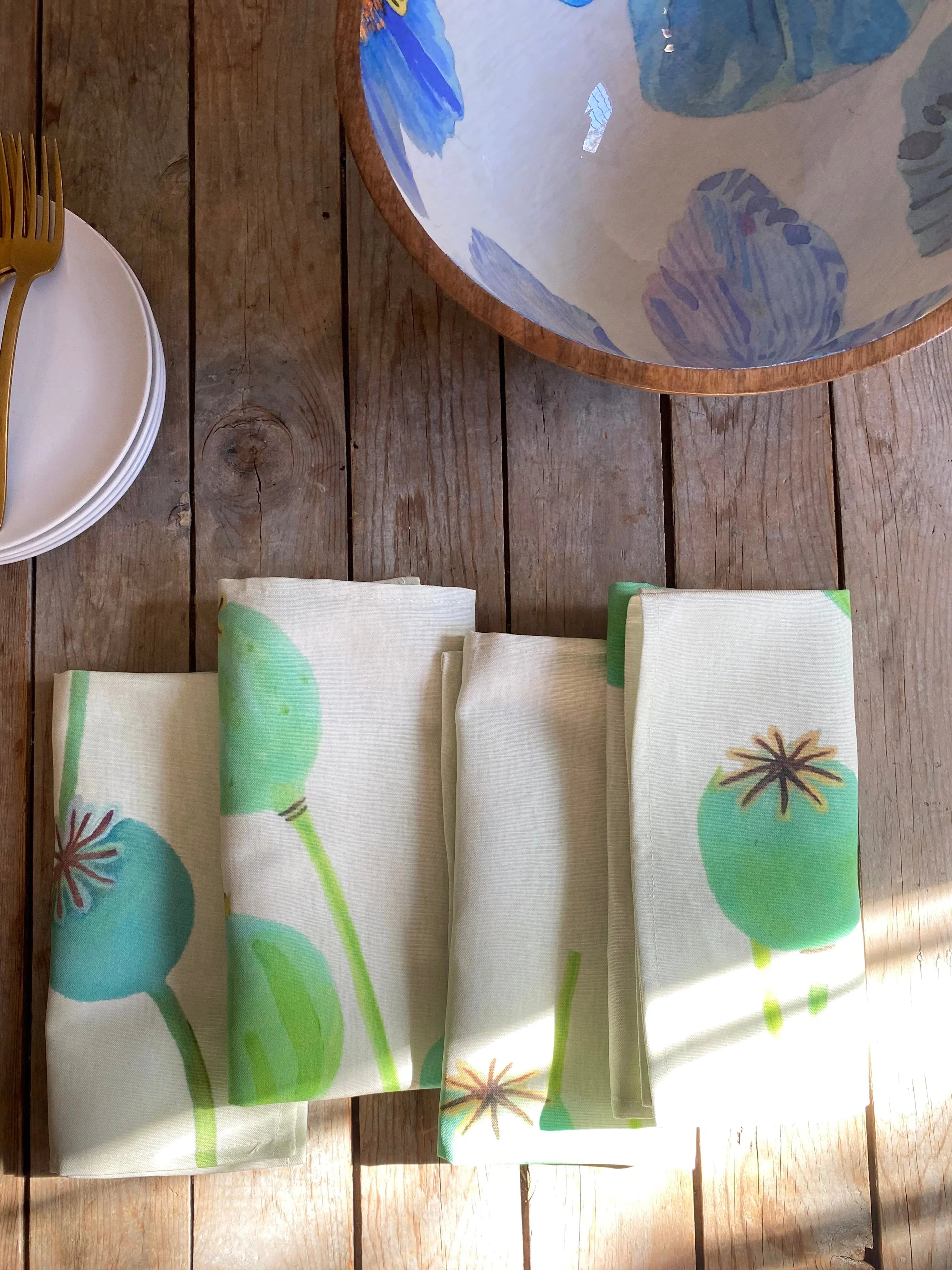 Folded ecru napkins with green poppy pod prints on rustic wood table with partial white plates and gold fork, blue floral bowl