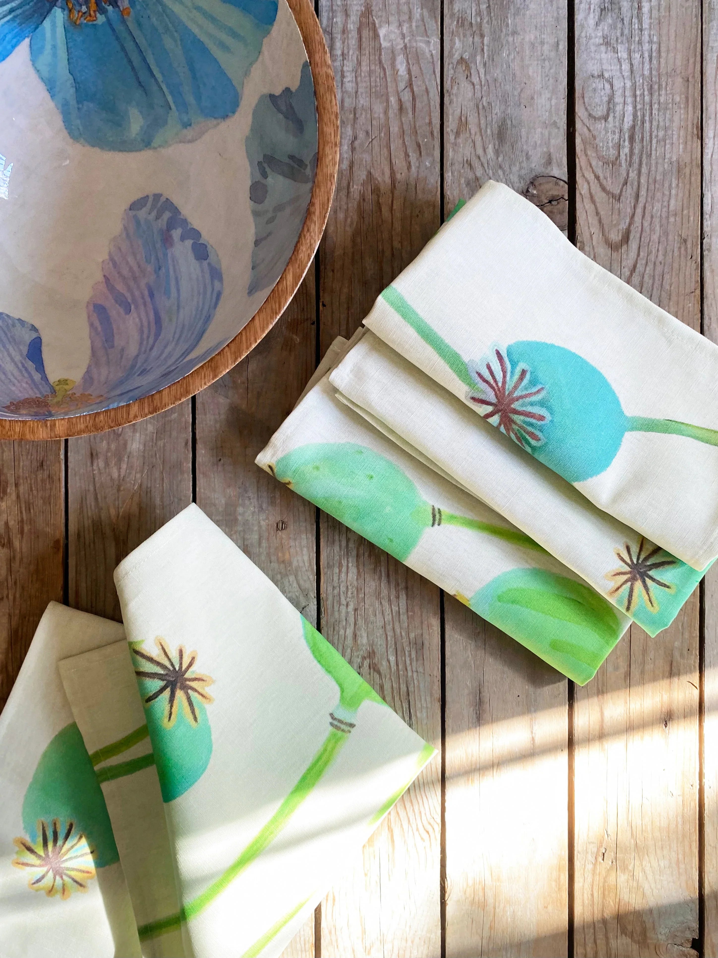 Folded cream napkins with green and blue poppy pod prints on wooden table next to floral bowl