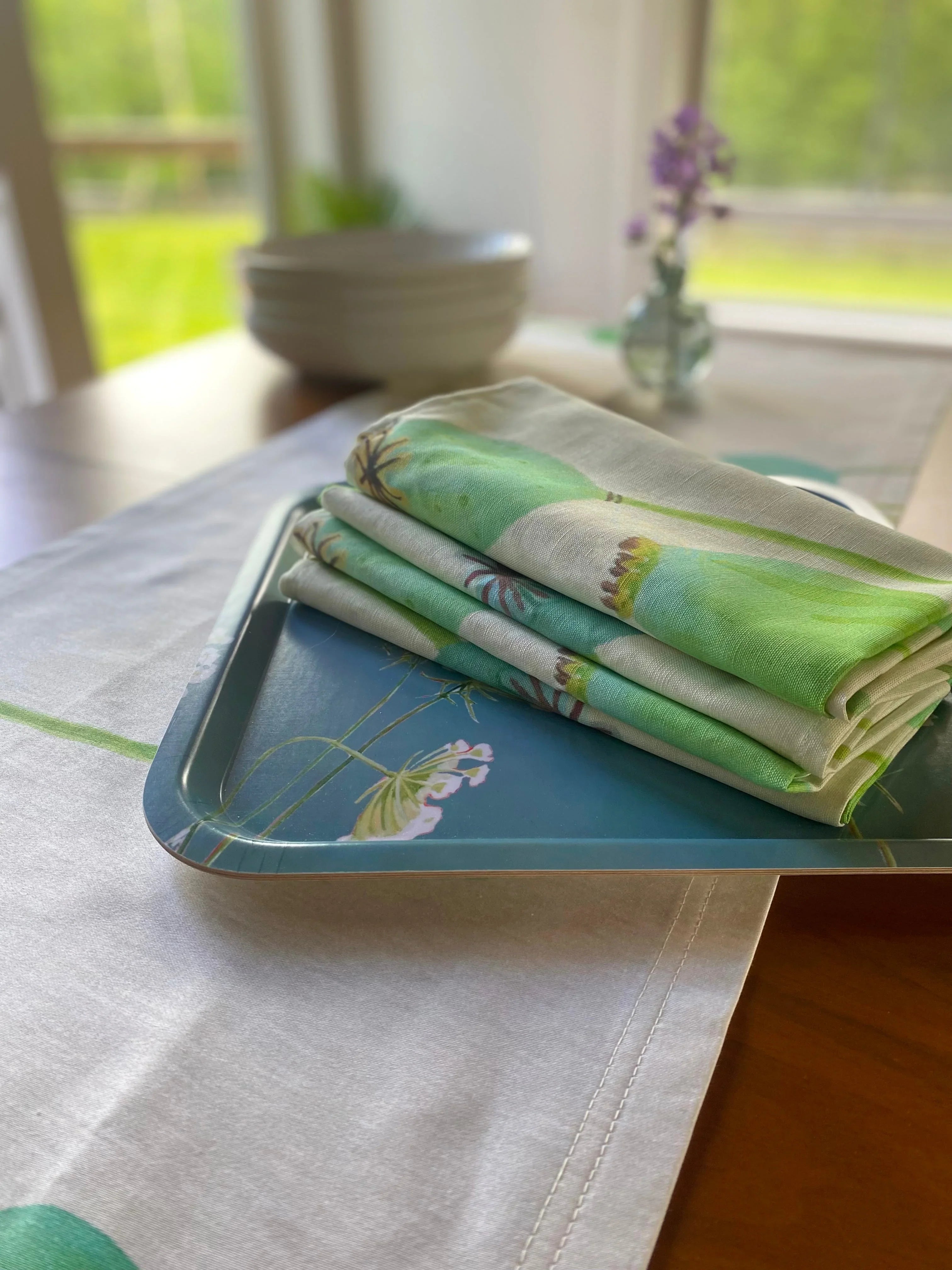 Folded green botanical napkins on blue floral tray with stacked white bowls and vase in background