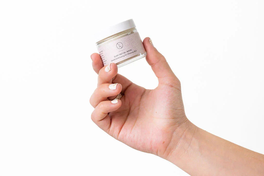 Hand holding a small jar of gentle clay facial mask for glowing skin against white background