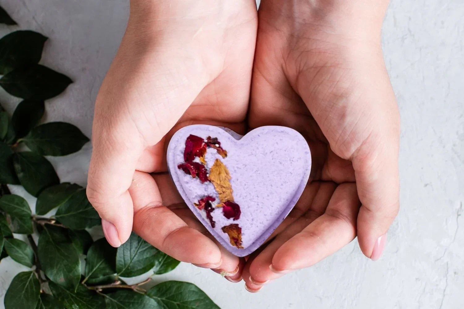 Hands holding a heart-shaped purple bath bomb with dried flower petals on a white surface with green leaves