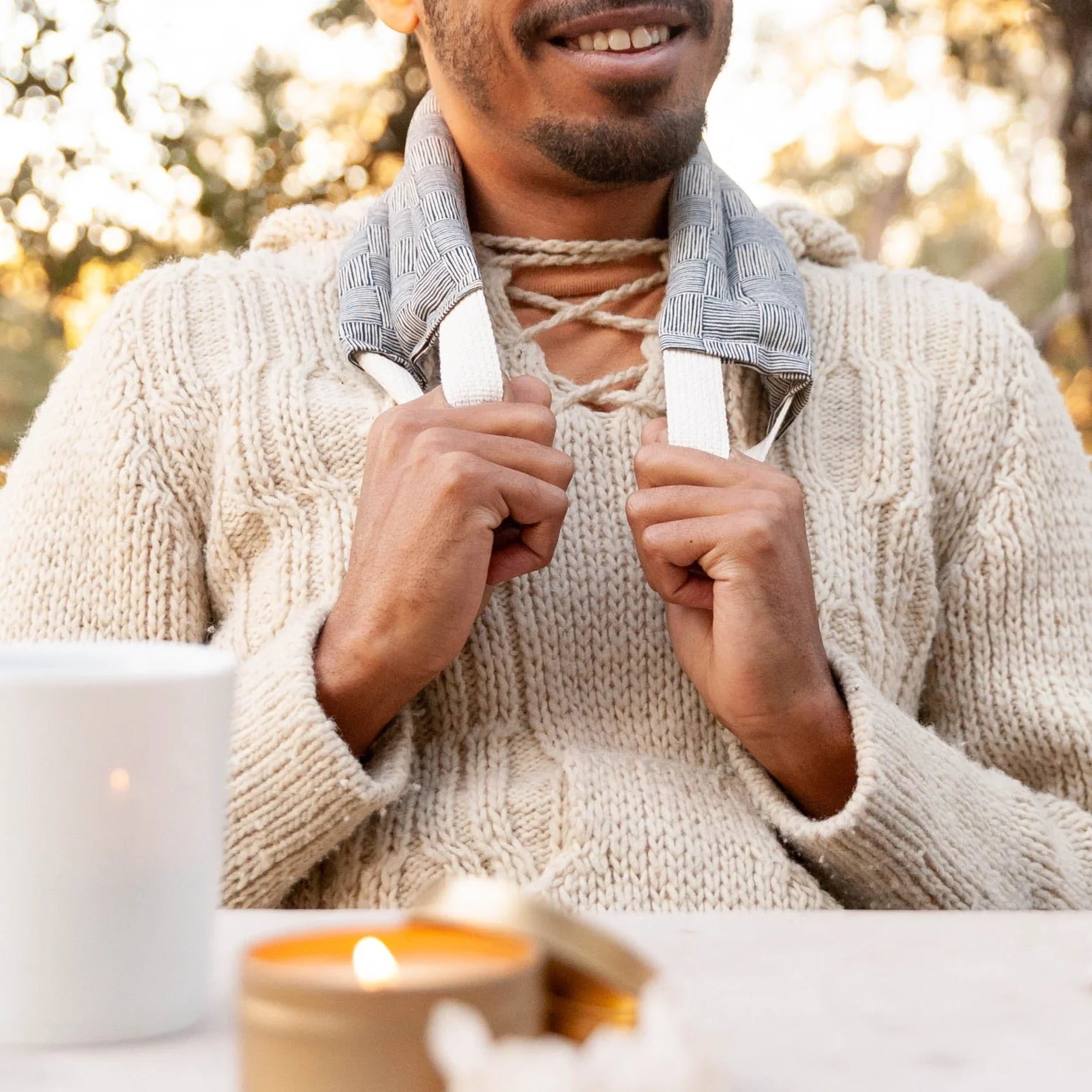 Man in beige chunky knit sweater using gray neck wrap outdoors with lit candles on table