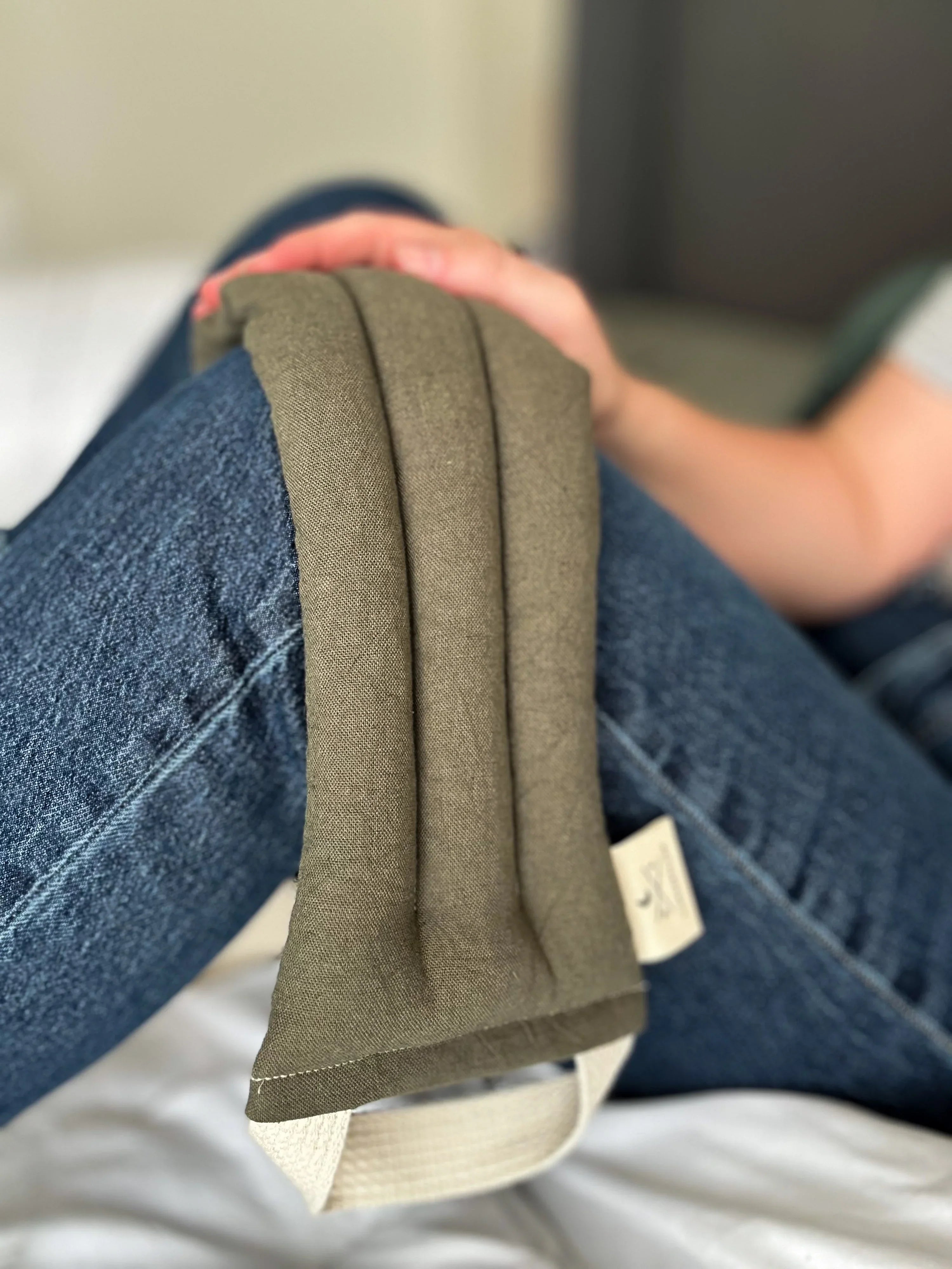 Person holding a greenish-brown therapy pack over bent knee wearing blue jeans on a white bed