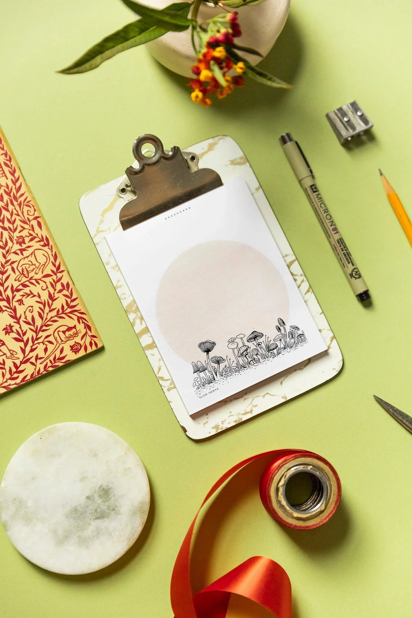 Notepad with wild mushroom illustration on clipboard surrounded by pen, sharpener, floral book, ribbon, and plant on green surface