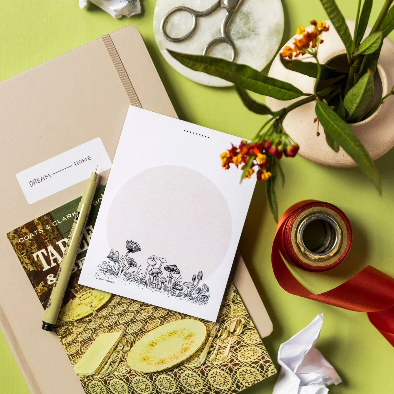 Notepad with wild mushroom illustration, beige journal, pen, vintage tableware book, scissors, red ribbon, and flowers on green surface