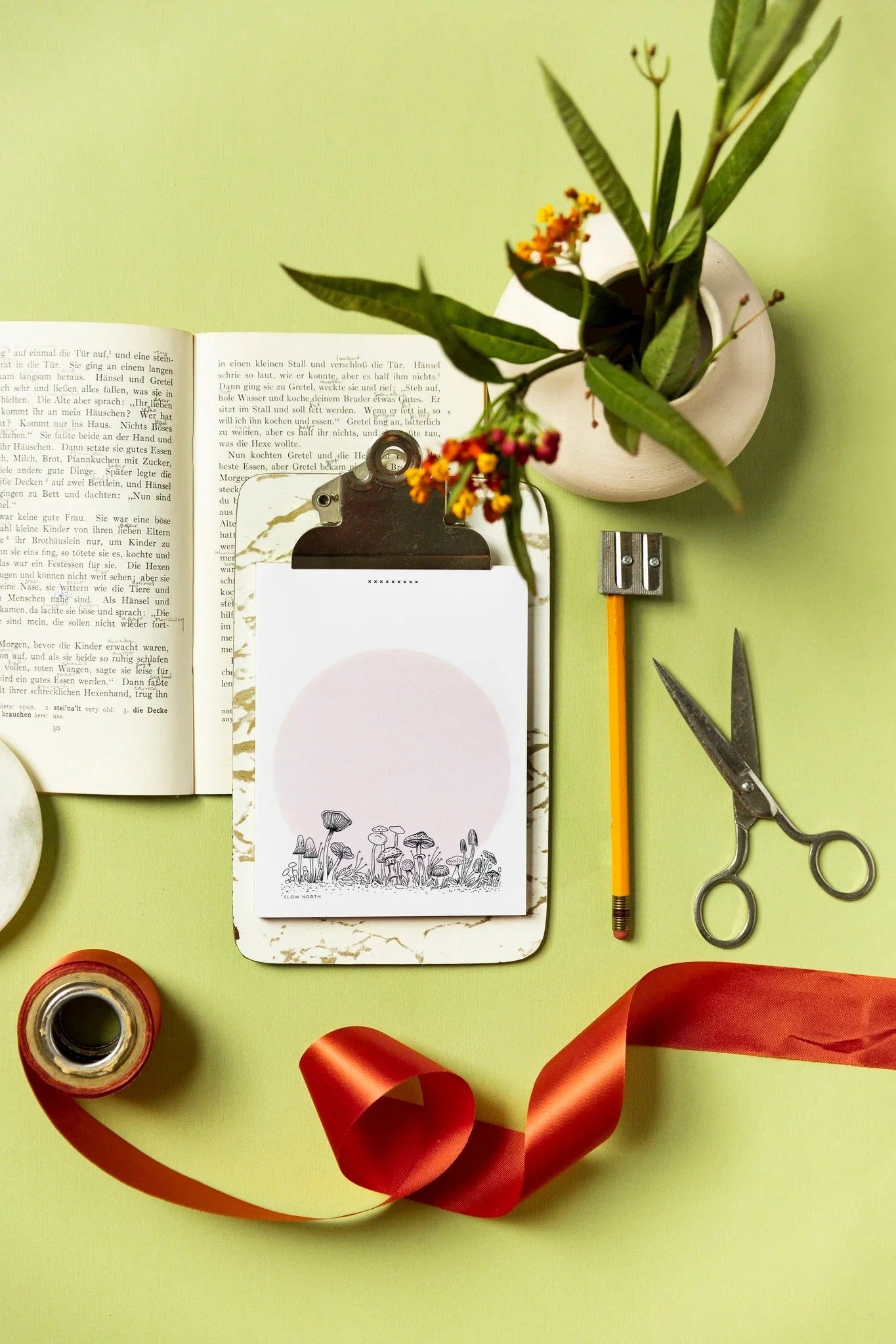 Flat lay of notepad with mushroom illustration on clipboard, scissors, pencil, red ribbon, and flower vase