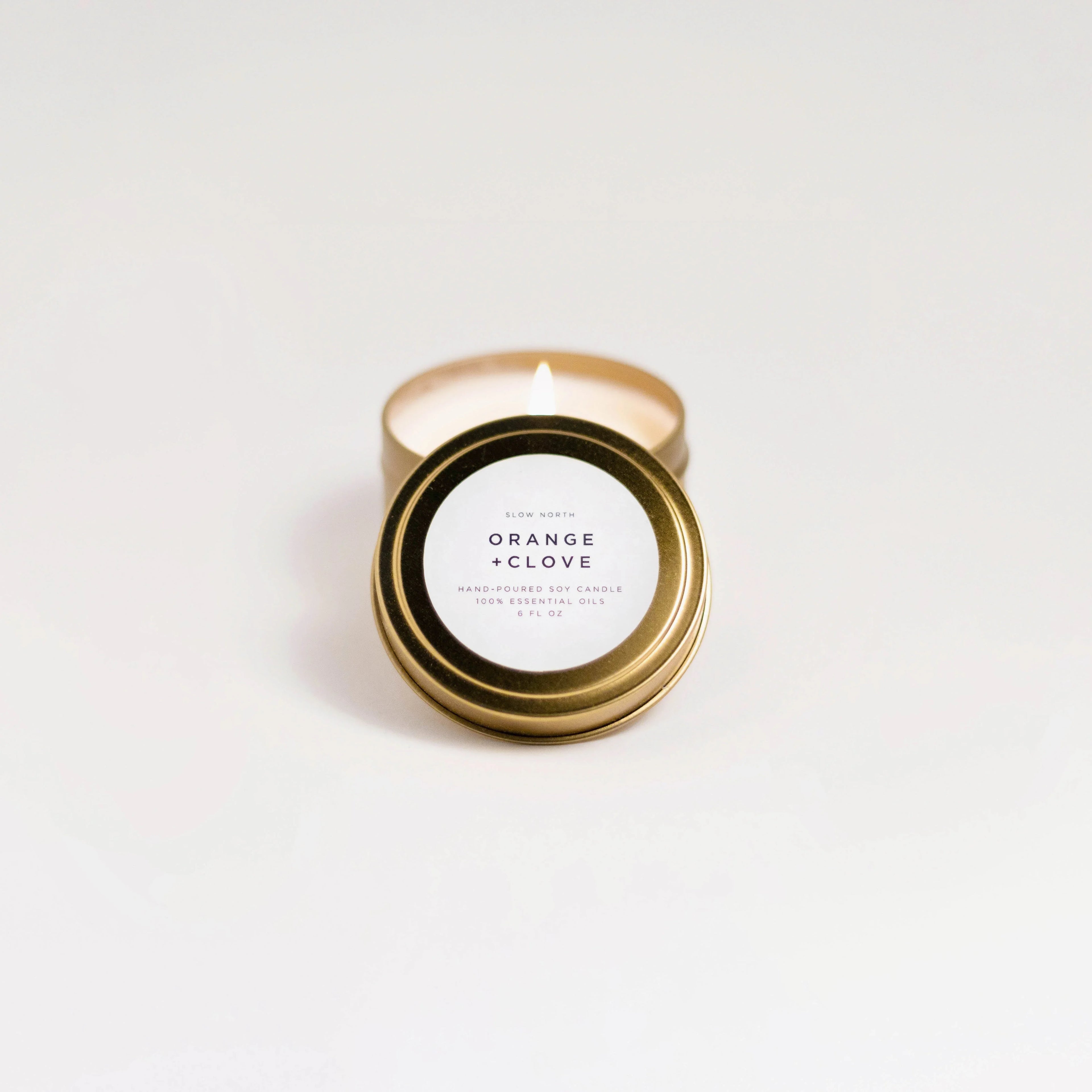 Lit orange and clove soy candle in gold travel tin with white label on white background