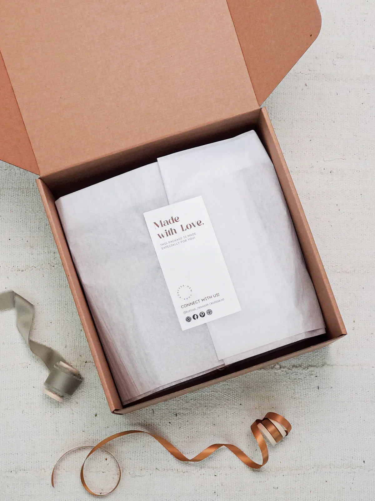 Brown gift box with white tissue paper and a card reading Made with Love, decorative ribbons nearby