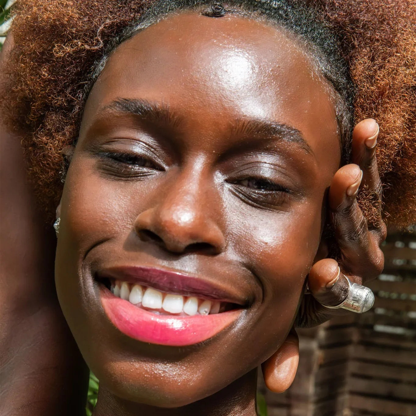 Smiling woman with natural hair and pink lipstick, wearing a silver ring, outdoors in sunlight