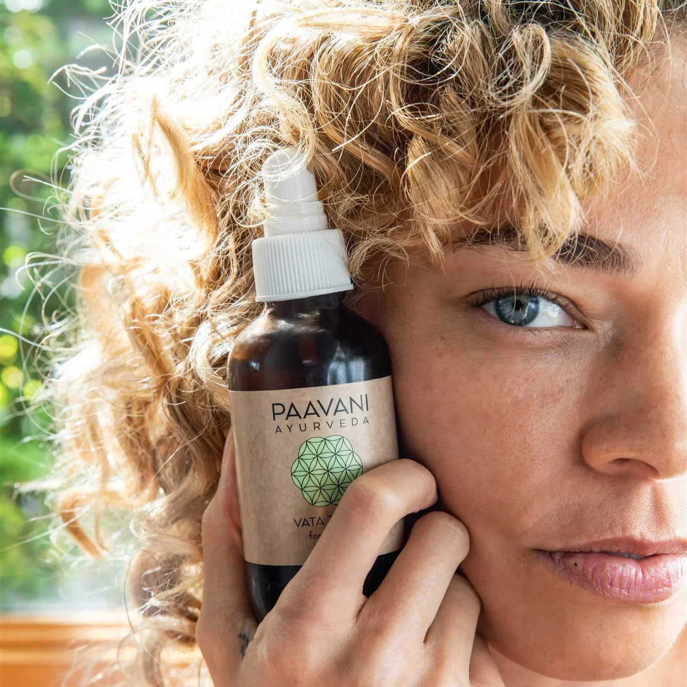 Woman with curly blonde hair holding Paavani Ayurveda skincare bottle near face, natural light