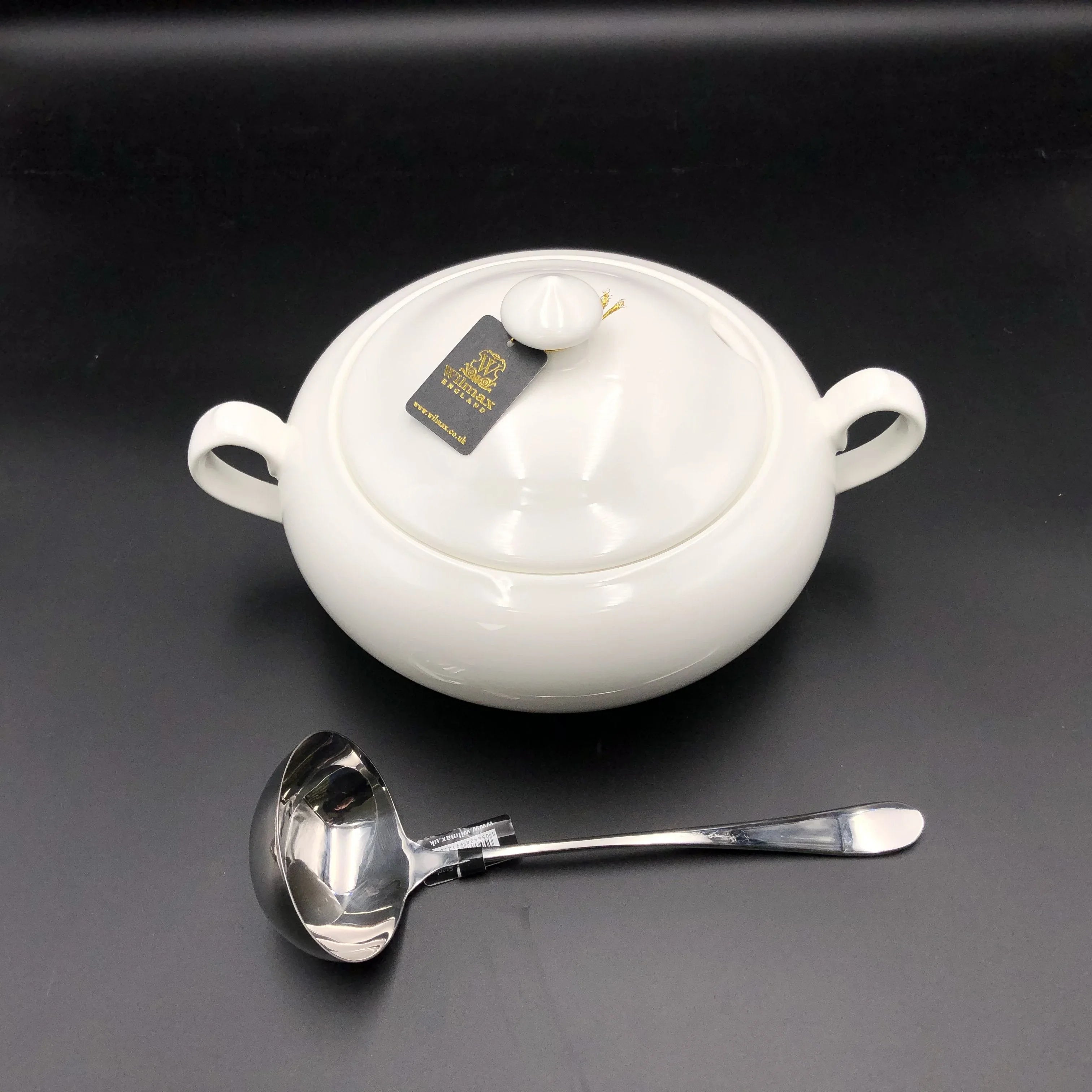Elegant white ceramic soup tureen with lid and handles beside a stainless steel ladle on black background