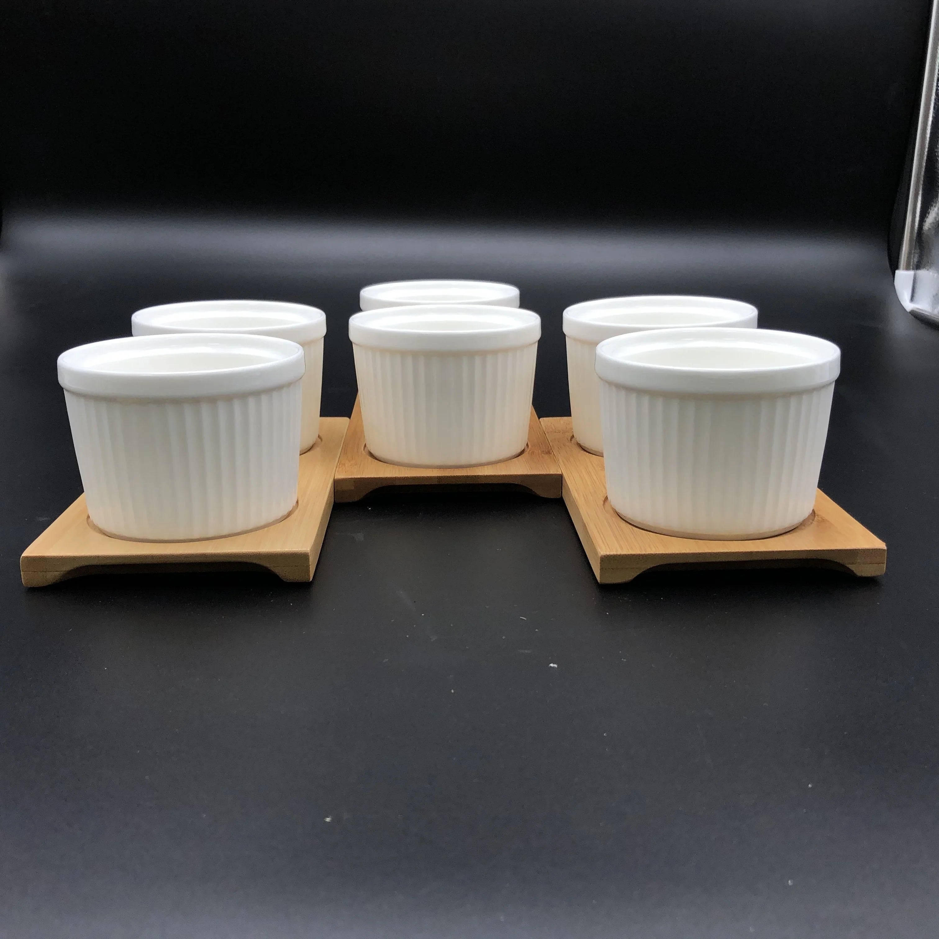 Set of six white ceramic ramekins on wooden trays, displayed on a black background