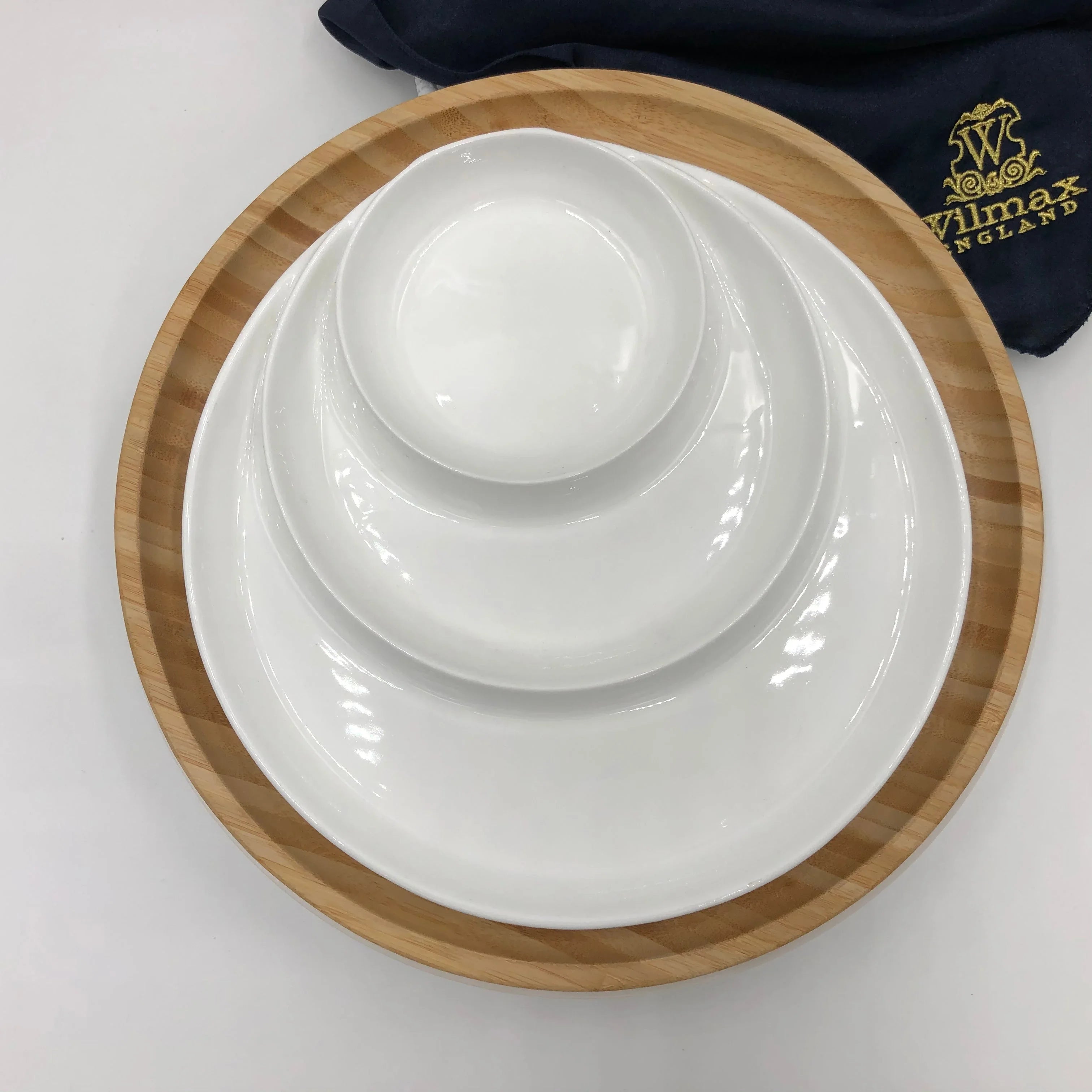 Stack of three white ceramic plates and bowl on round wooden tray with dark blue Wilmax England cloth