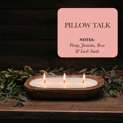 Pillow Talk soy candle in rustic wooden bowl with three flames, greenery, floral scent notes.