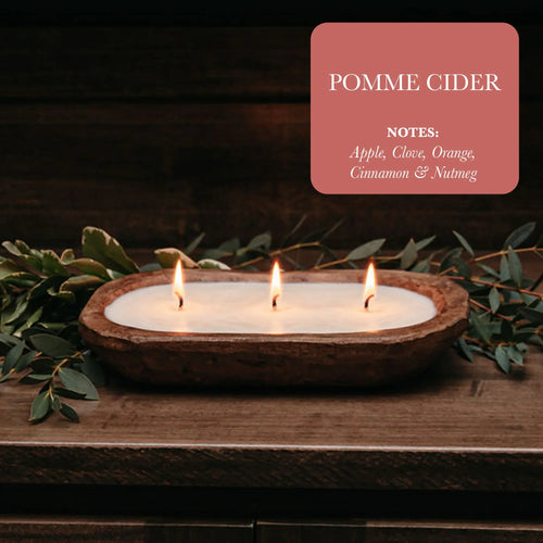 Pomme Cider scented candle with three flames in a rustic wooden bowl, surrounded by greenery on a wood table, highlighting notes of apple, clove, orange, cinnamon, and nutmeg from Qu!rk Boutique.