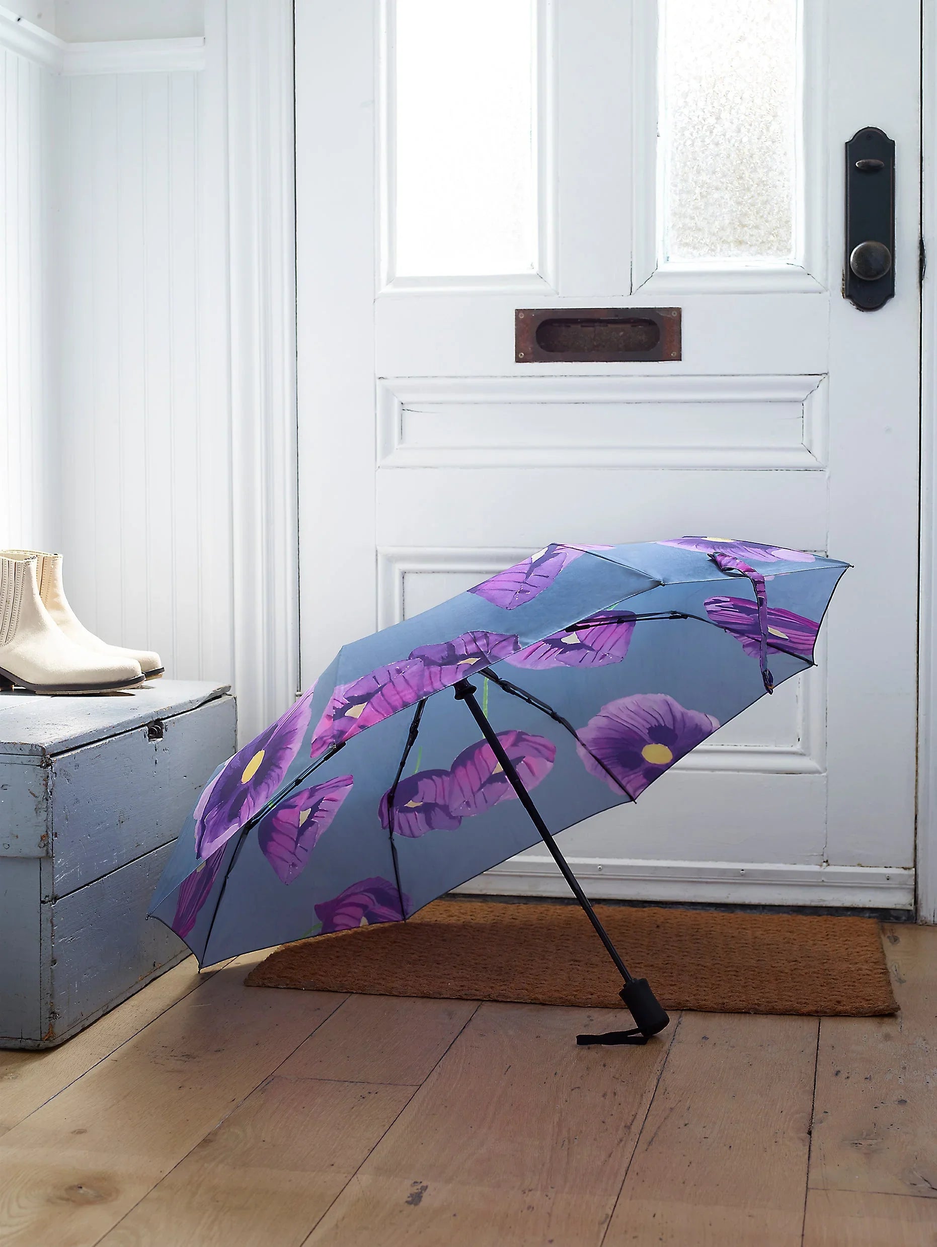 Open blue umbrella with purple poppy flower print leaning against white door on wooden floor