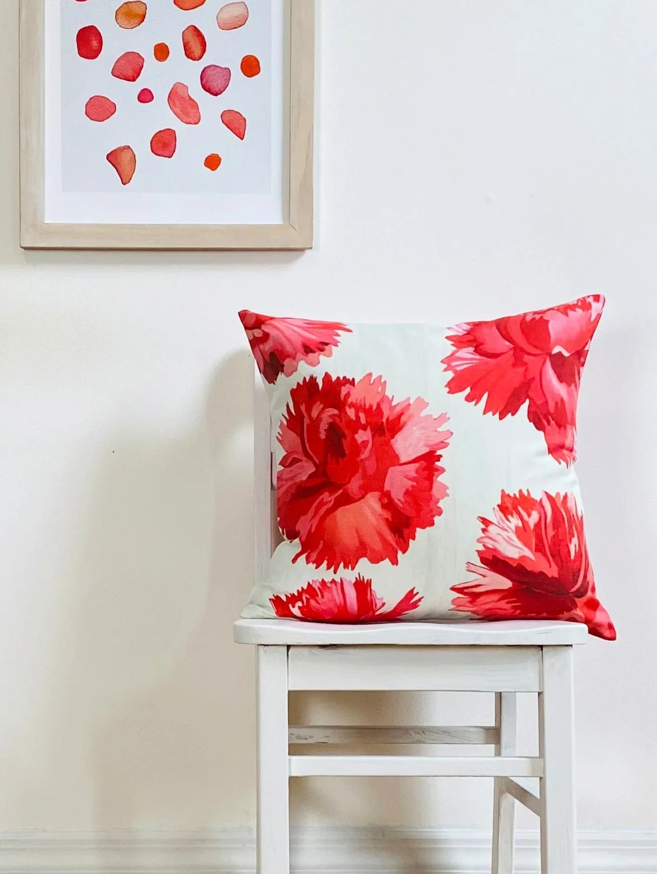 White chair with decorative pillow featuring large red carnation floral print against plain wall