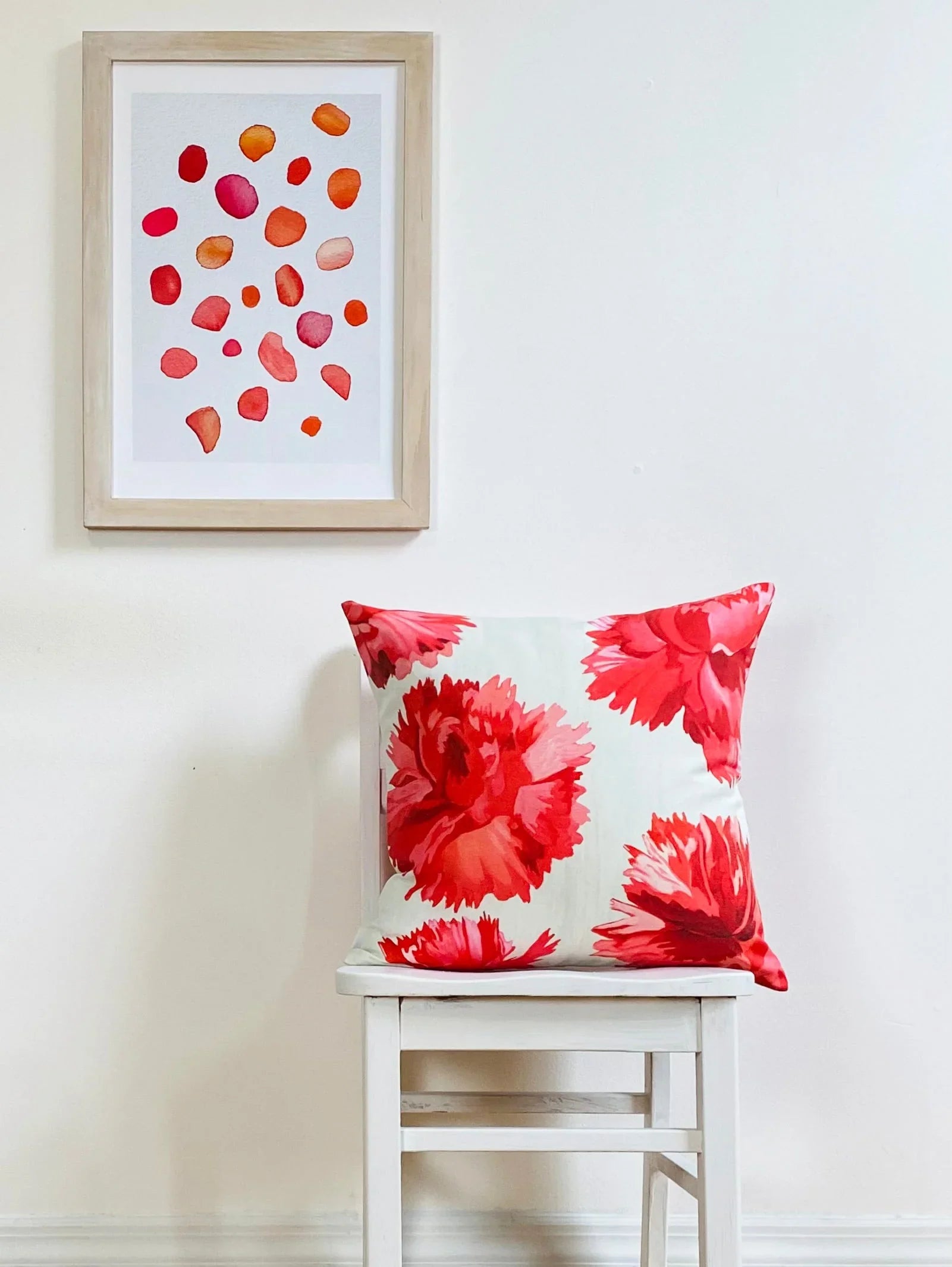White chair with a decorative pillow featuring large red carnation flowers and a framed artwork of scattered rose petals on the wall