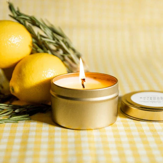 Lit rosemary lemon scented candle in gold tin with fresh lemons and rosemary on yellow checkered cloth