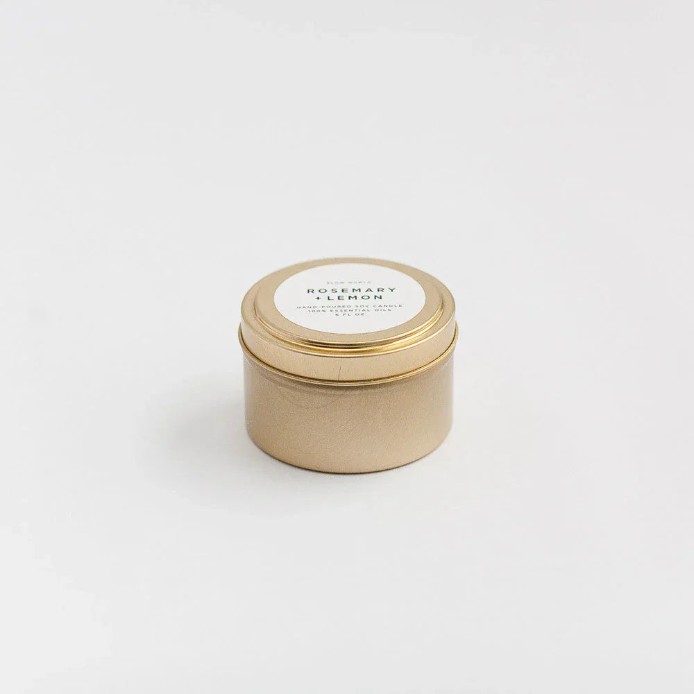Gold travel tin candle labeled Rosemary + Lemon on white background