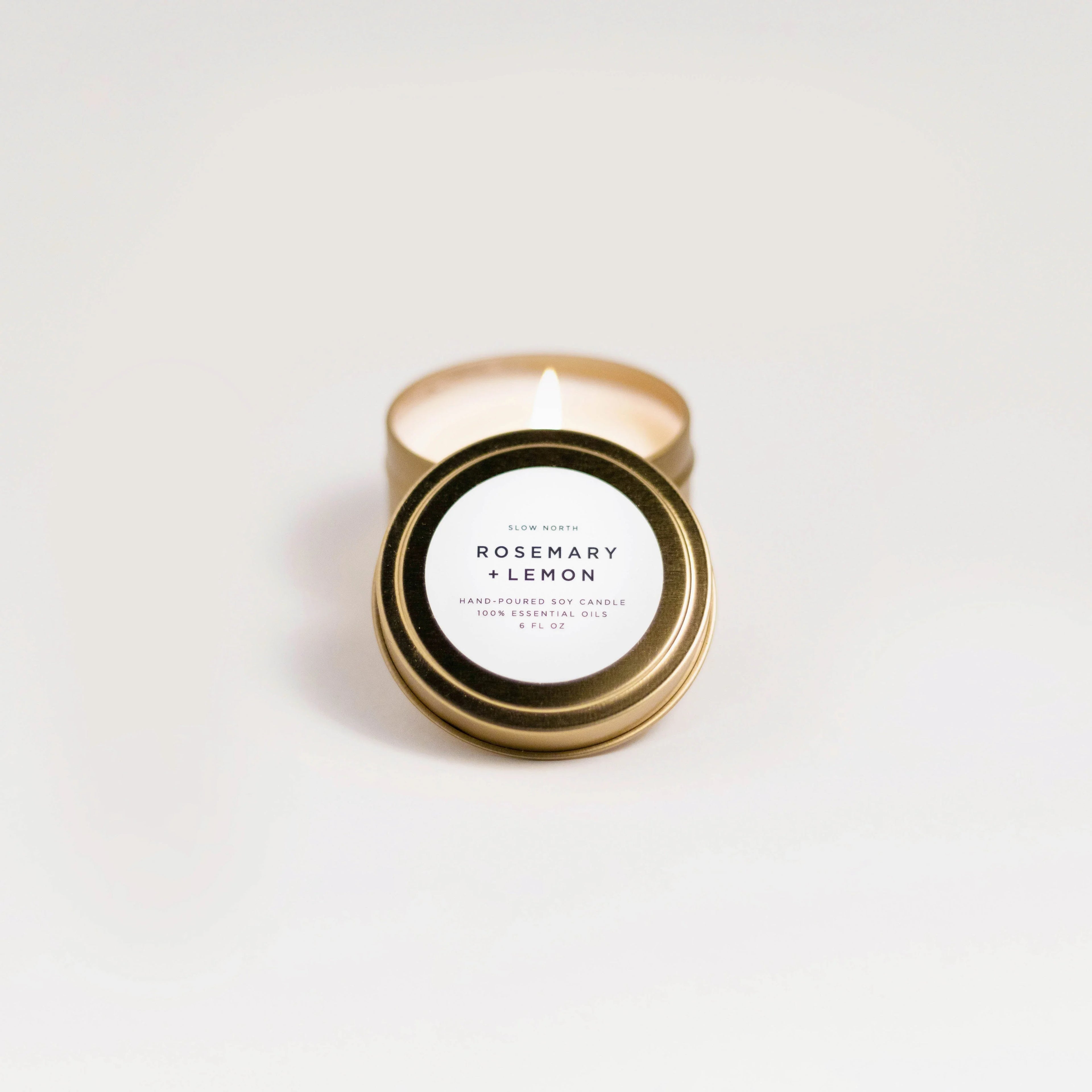 Lit hand-poured rosemary and lemon soy travel candle in gold tin on white background