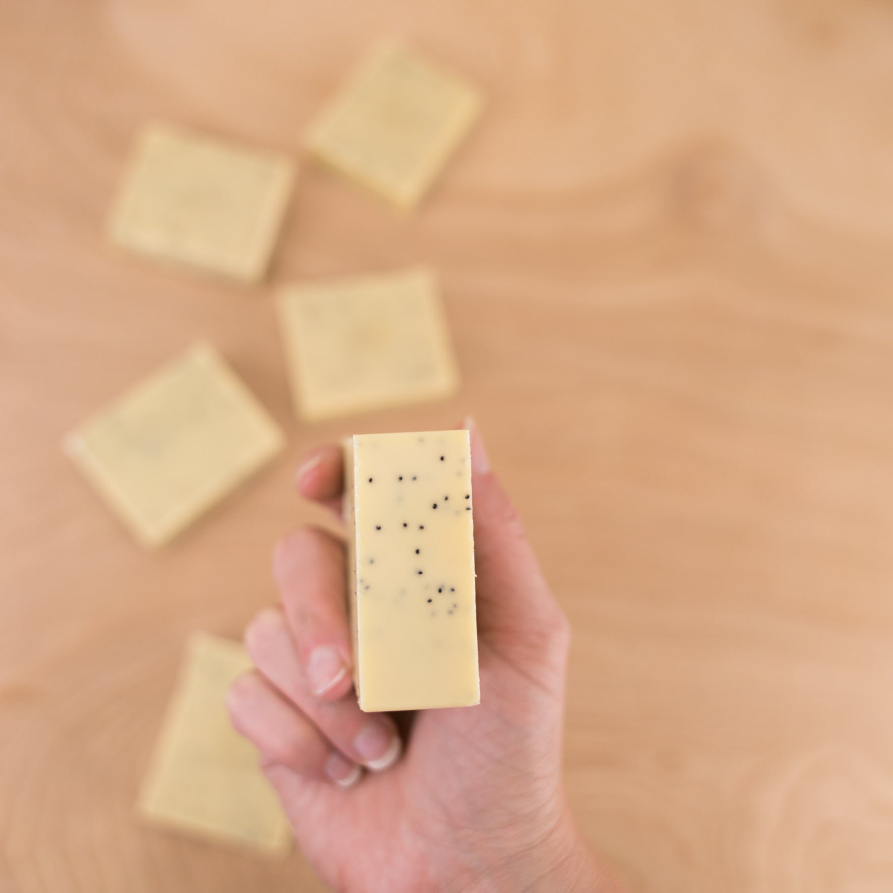 Hand holding rectangular yellow bar soap with black specks, other soap bars blurred on wooden surface