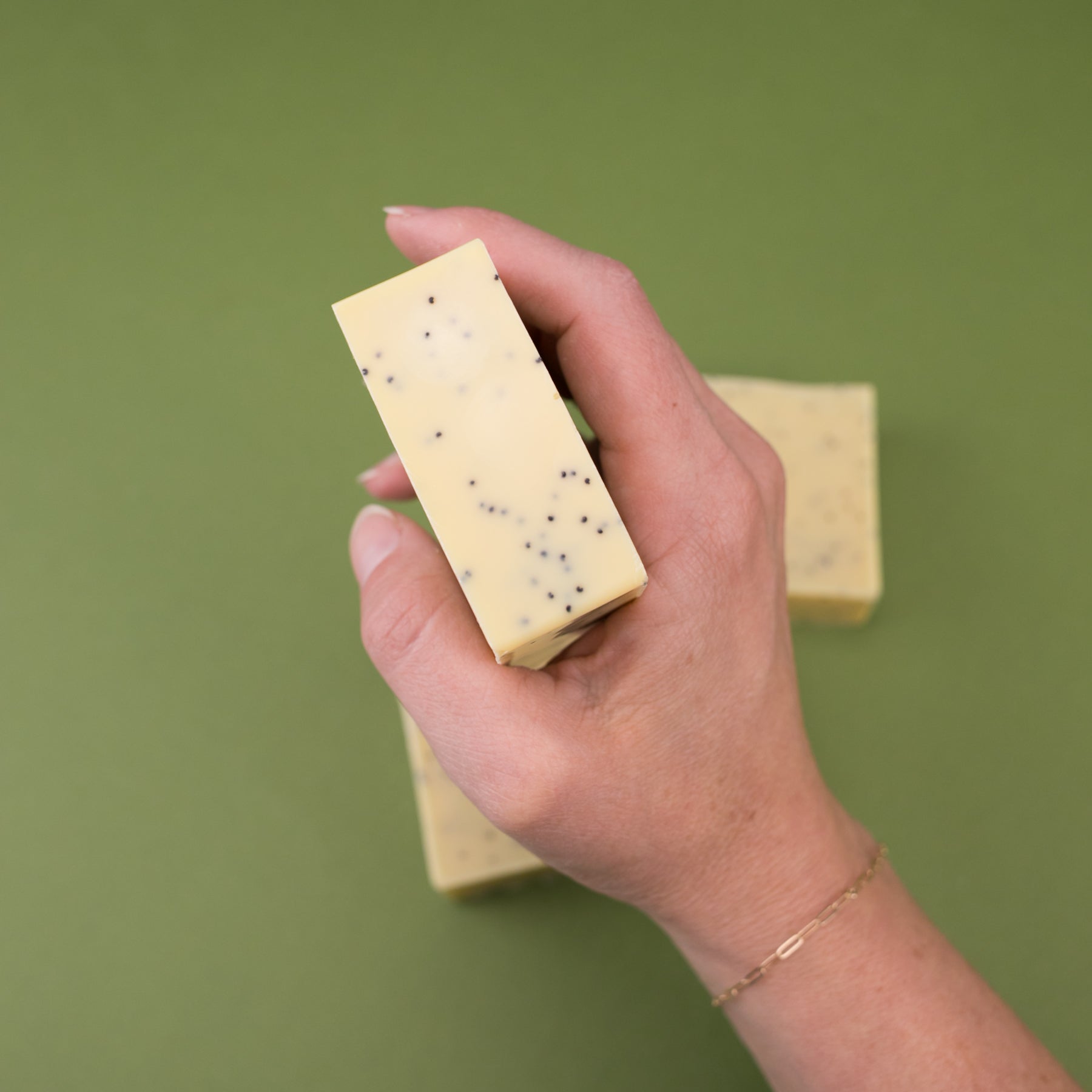 Hand holding a rectangular yellow rosemary lemon bar soap with small black specks on green background