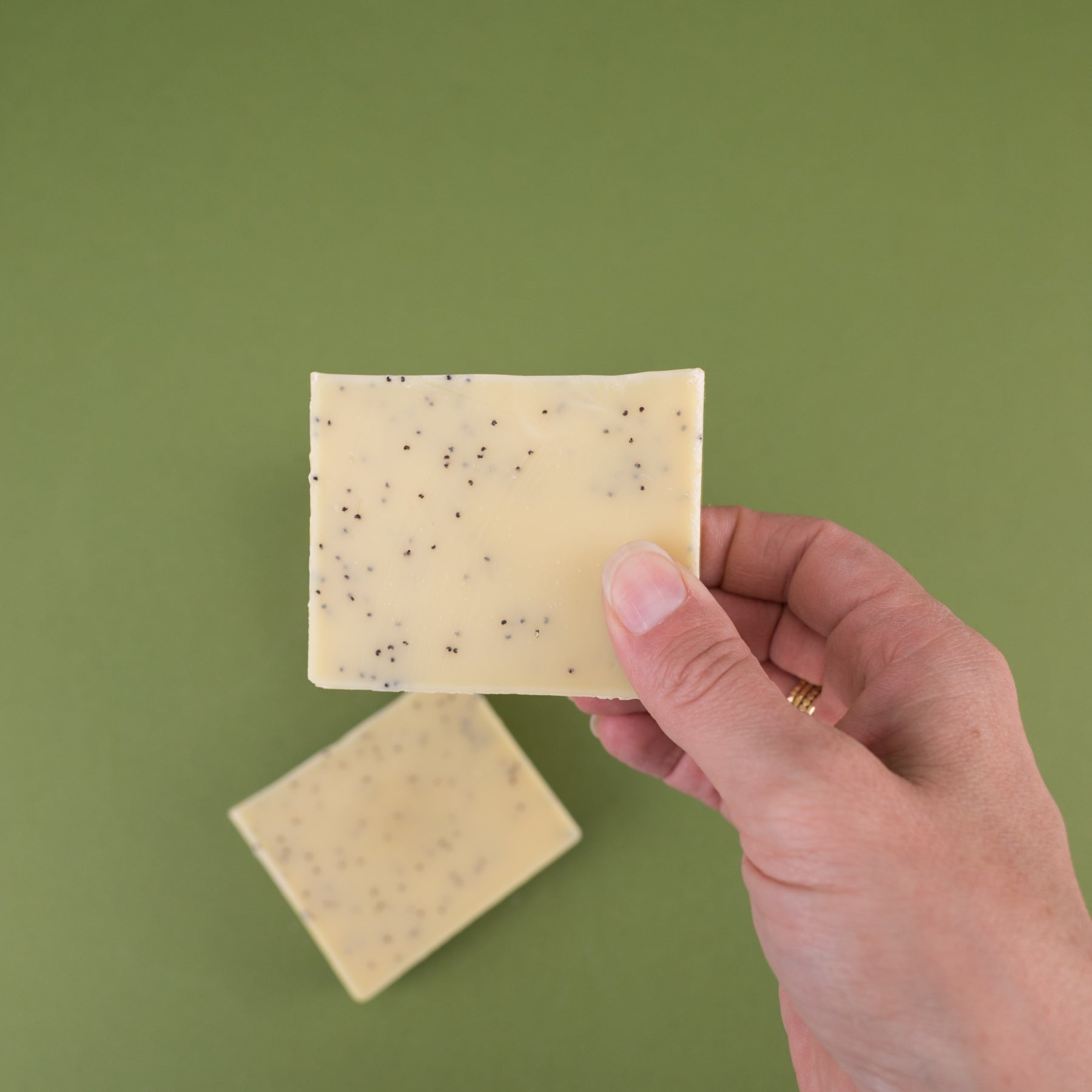 Hand holding a rectangular bar of yellow rosemary lemon soap with black specks on green background