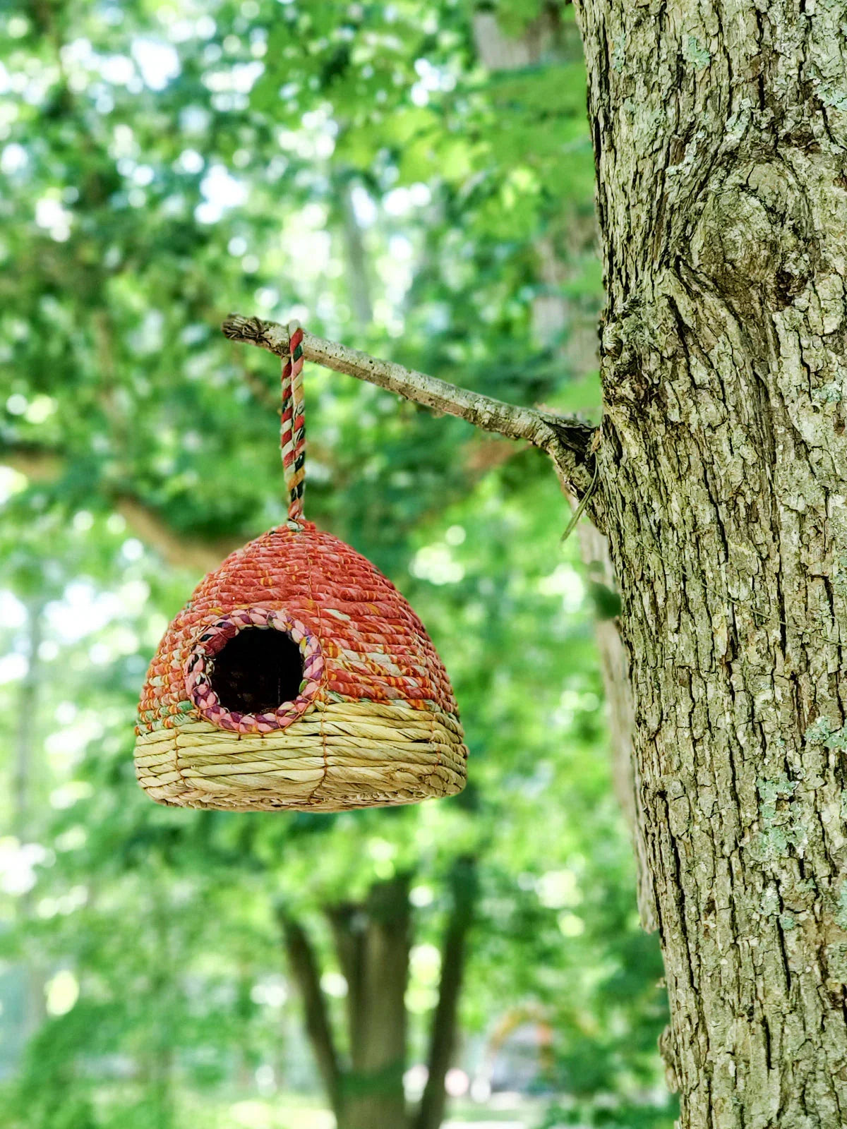 Handwoven natural straw birdhouse hanging from a tree branch in a lush green garden