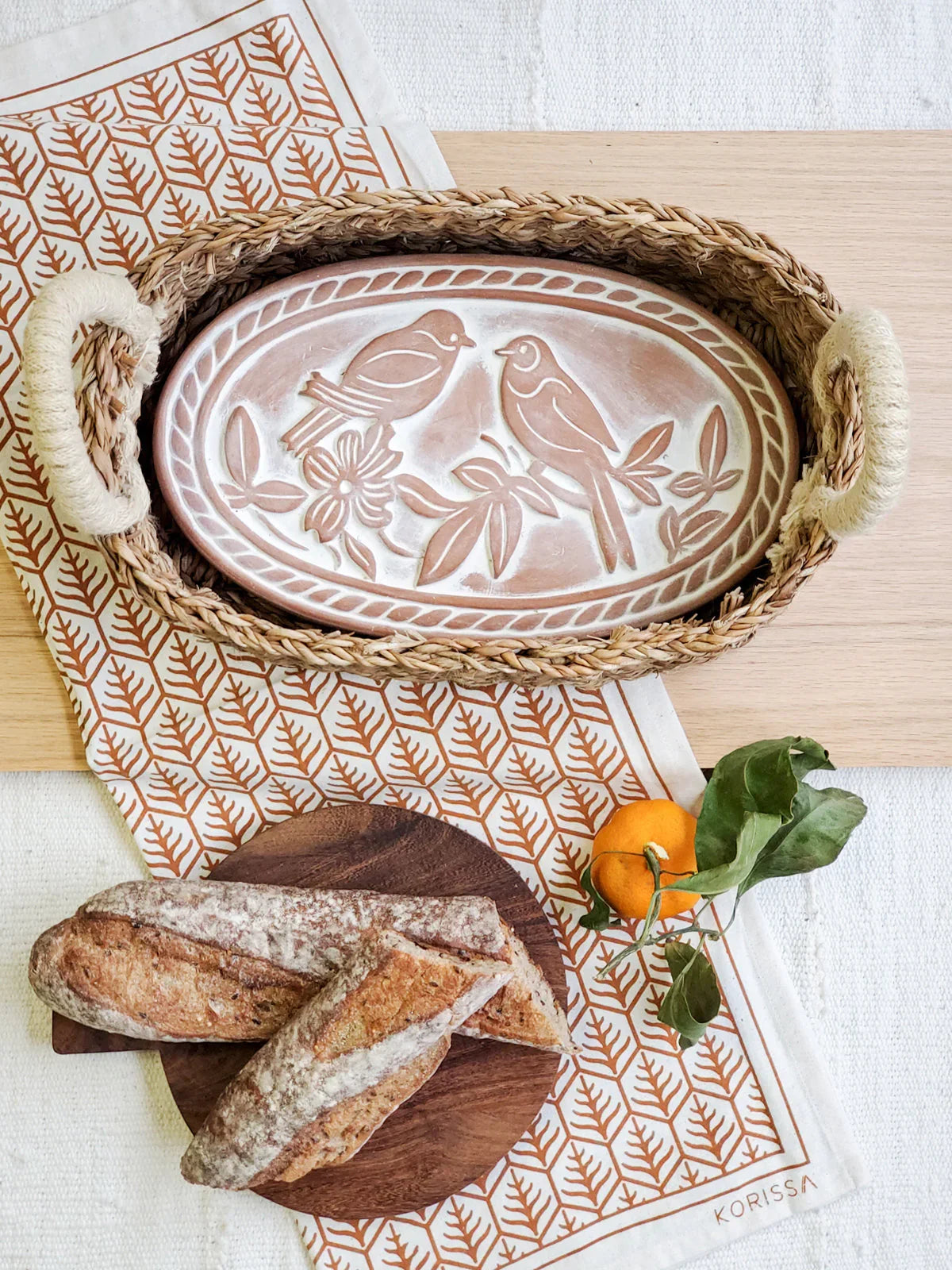 Oval woven bread basket with lovebird design lid, rustic baguettes on wooden board, orange fruit on patterned cloth
