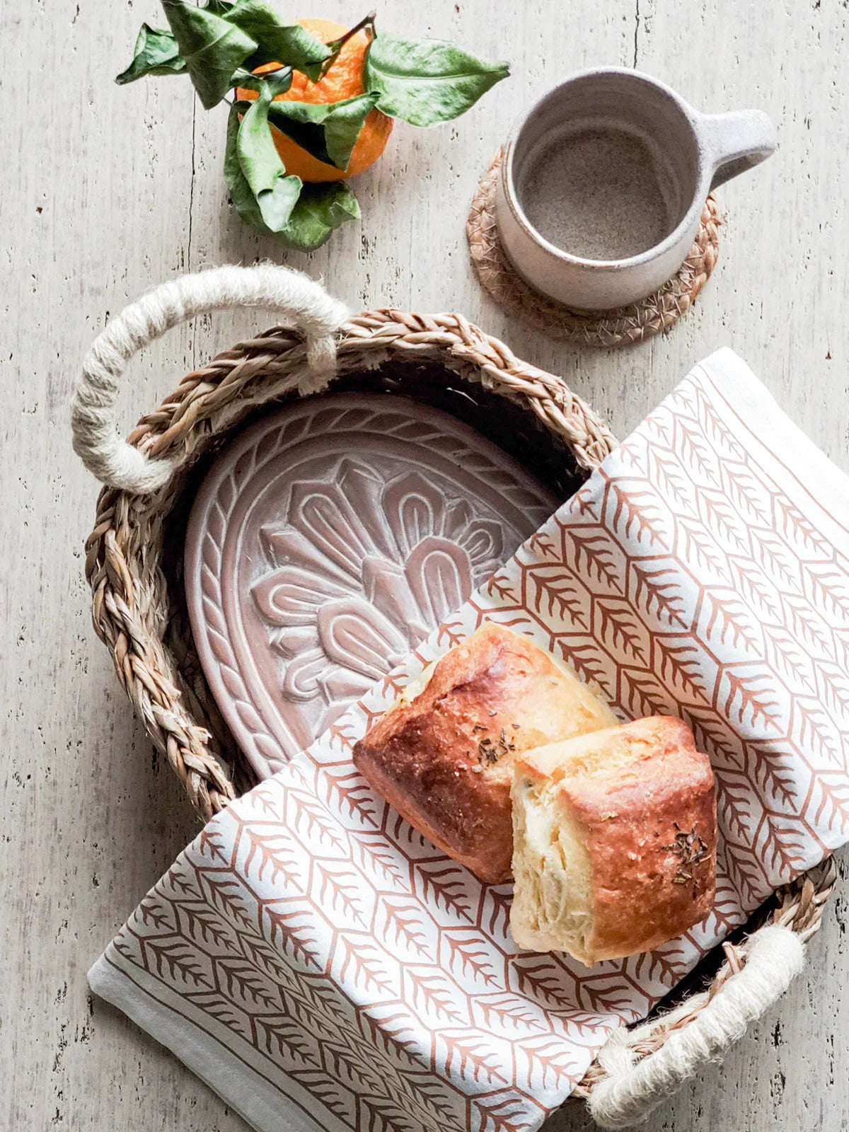 Woven basket with patterned tea towel holding two herb-topped bread rolls, clay plate and ceramic cup on rustic white table