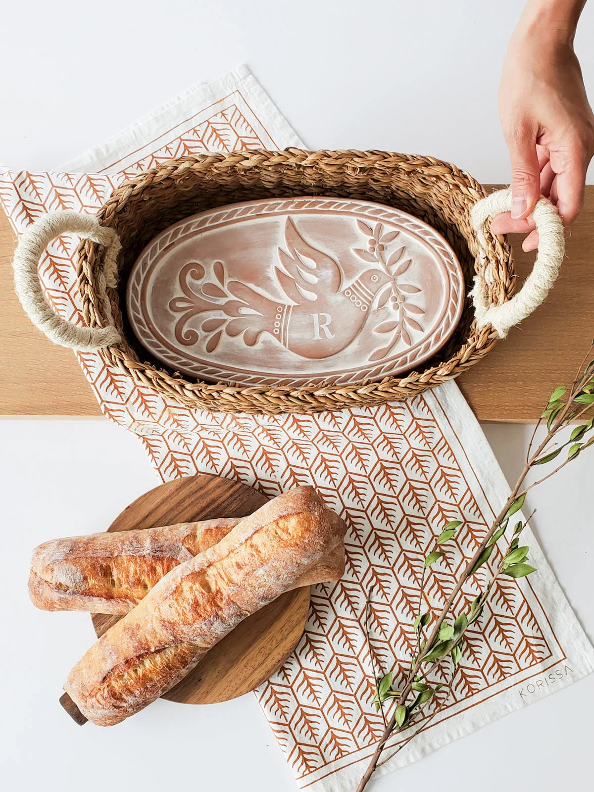 Hand holding woven bread basket with personalized embossed clay bread warmer on patterned cloth, fresh baguettes on wooden board