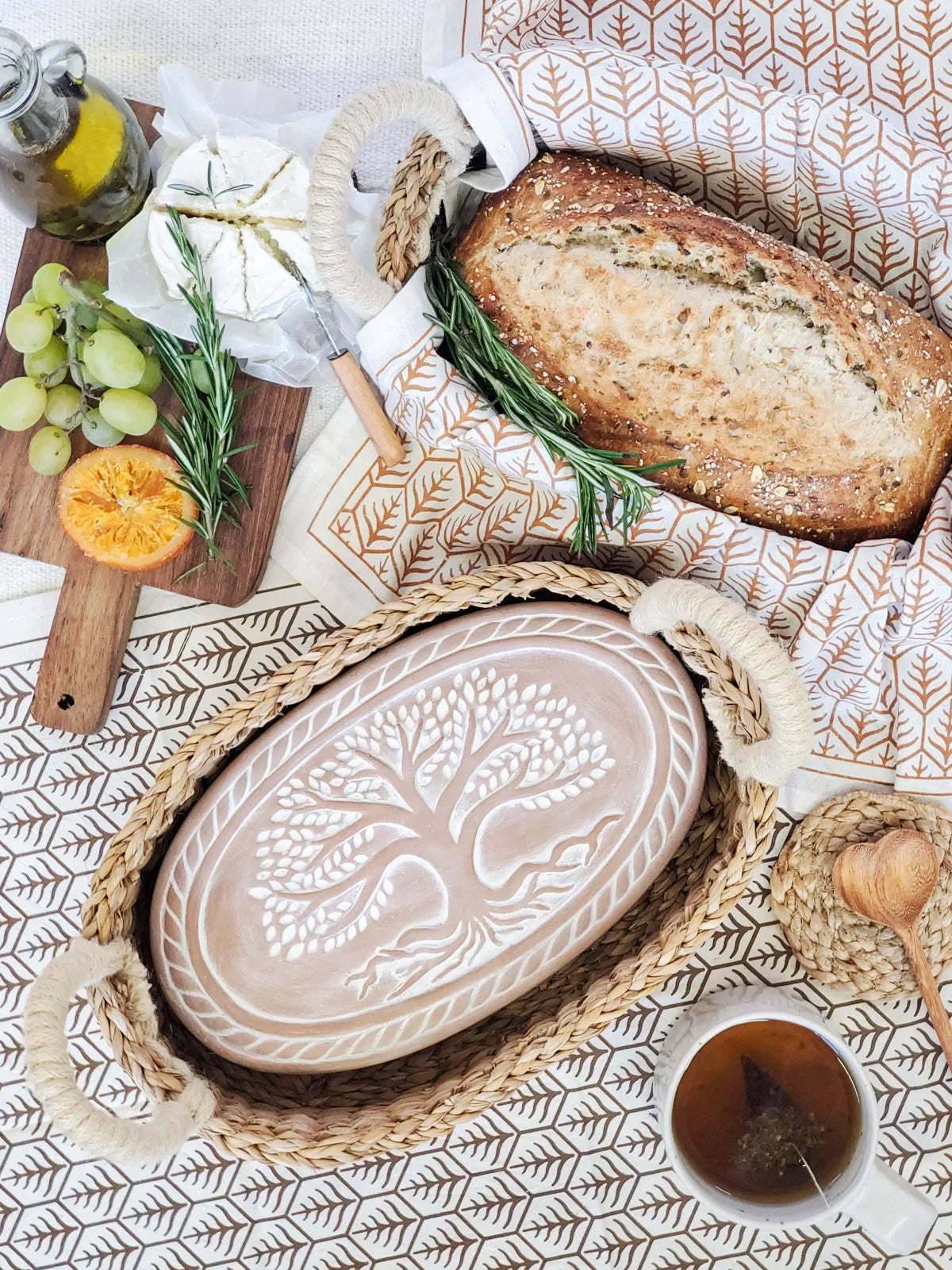 Woven bread warmer basket with carved Tree of Life bread baking dish, fresh bread, cheese, grapes, and herbal tea