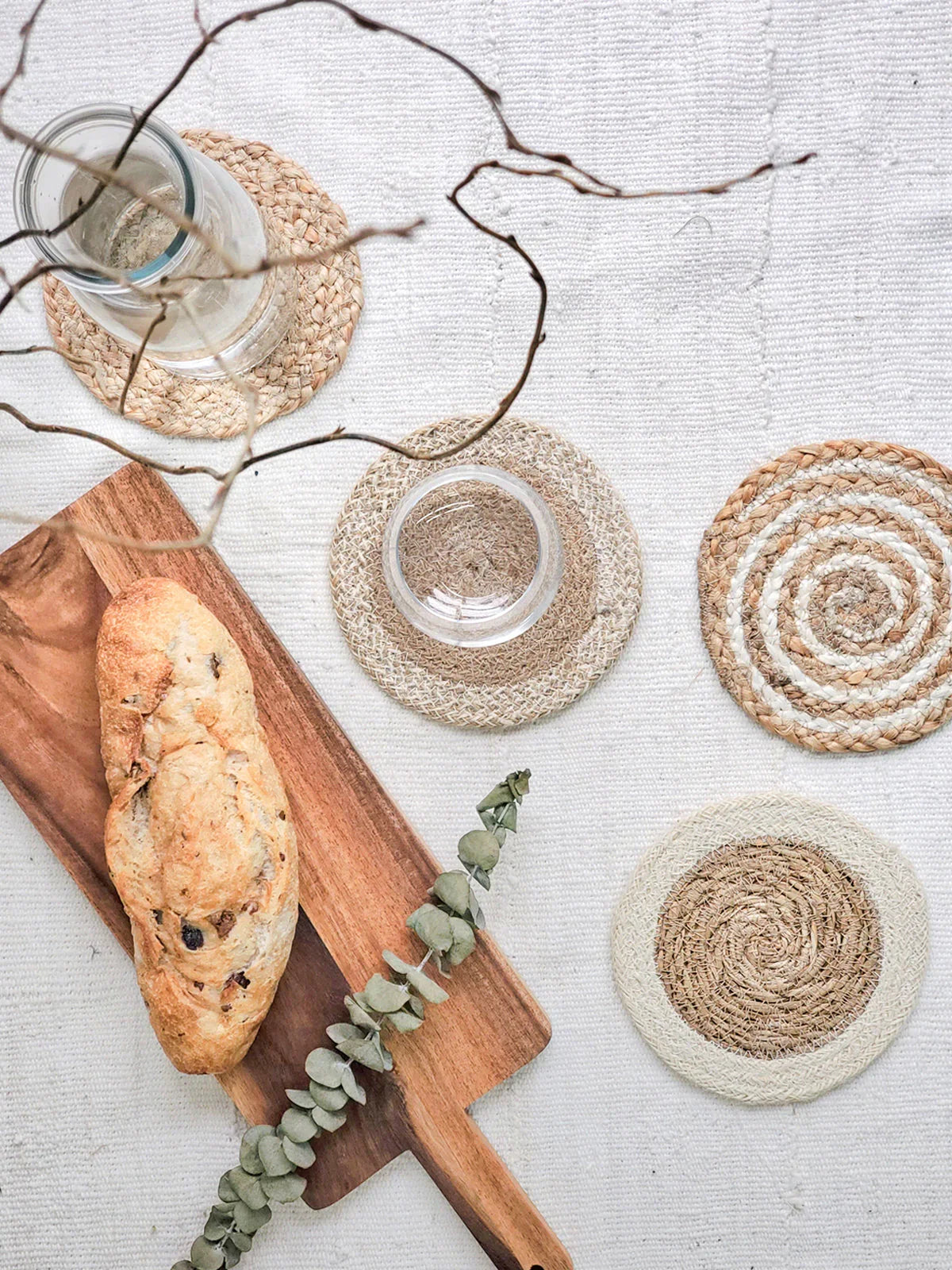 Rustic bread on wooden board with glass on woven coaster and round jute trivets on white linen