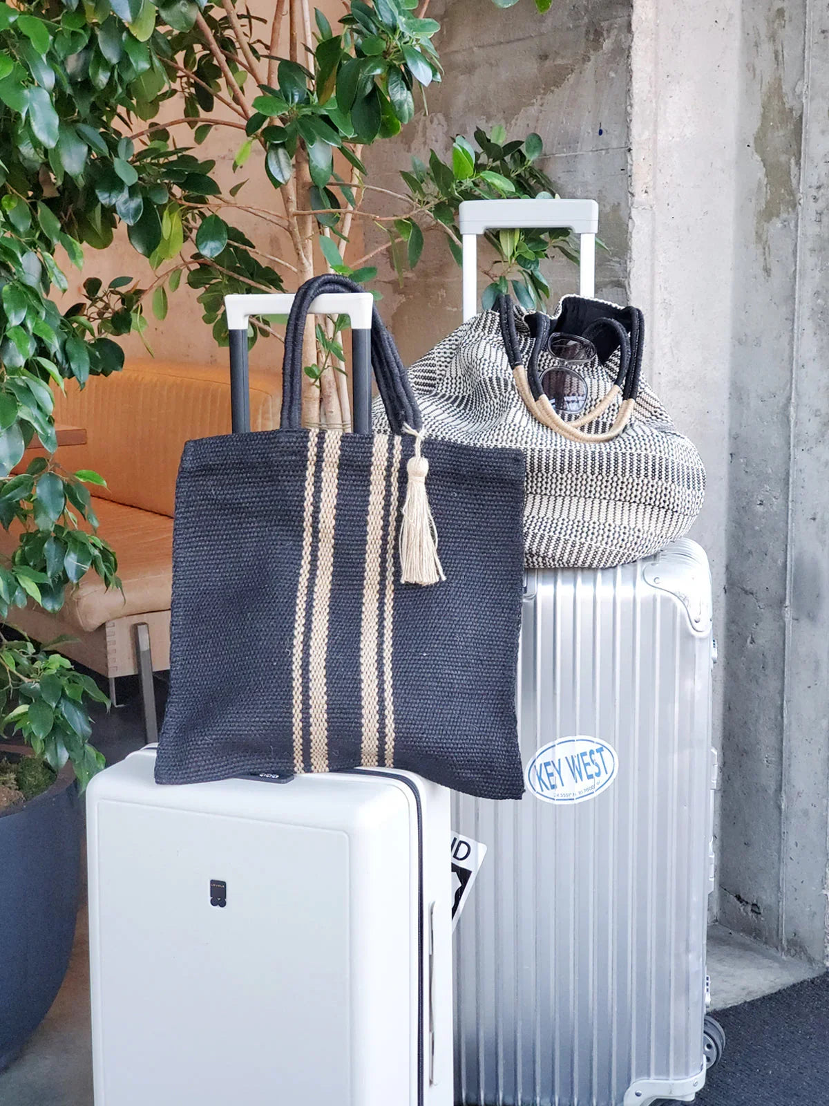 Two modern suitcases with black and patterned woven tote bags, indoor setting with plants and concrete wall