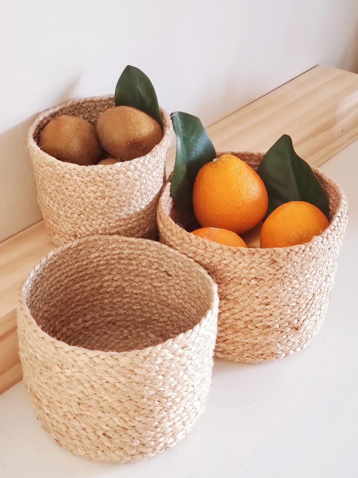 Three natural woven baskets on wooden shelf, two filled with kiwis and oranges with green leaves