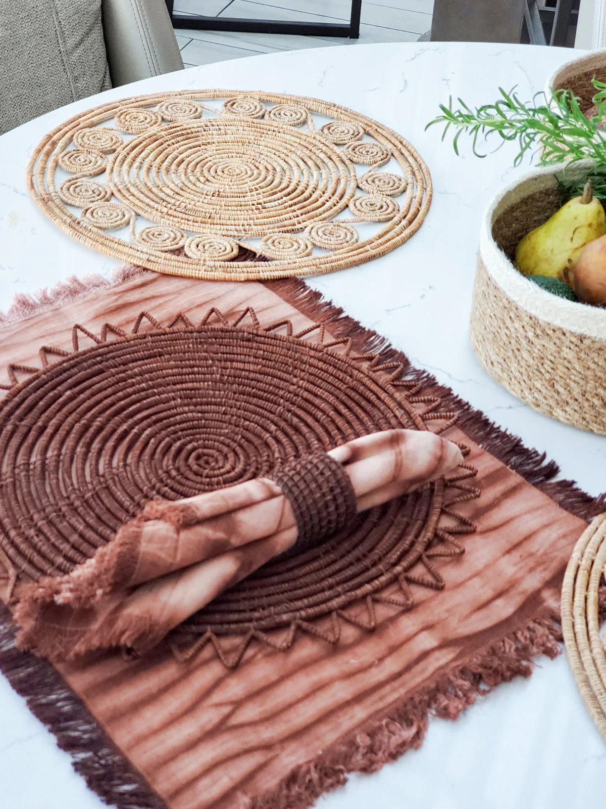 Brown woven placemat set on white marble table with frayed cloth napkin and basket of fruit