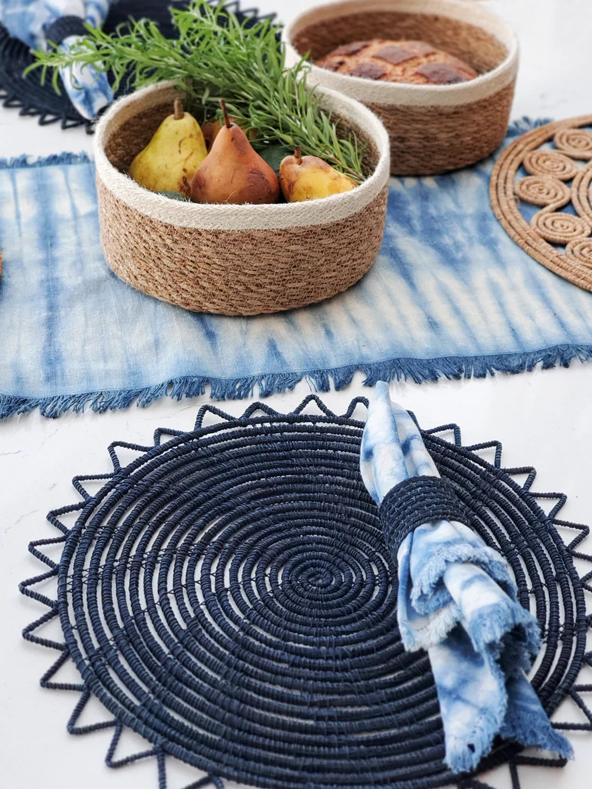 Navy woven circular placemat with blue tie-dye cloth napkin on white table, natural fiber baskets with pears and herbs