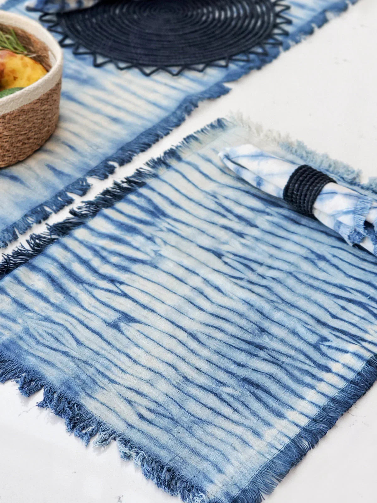 Blue and white tie-dye cotton placemat with fringed edges and matching rolled napkin on white surface