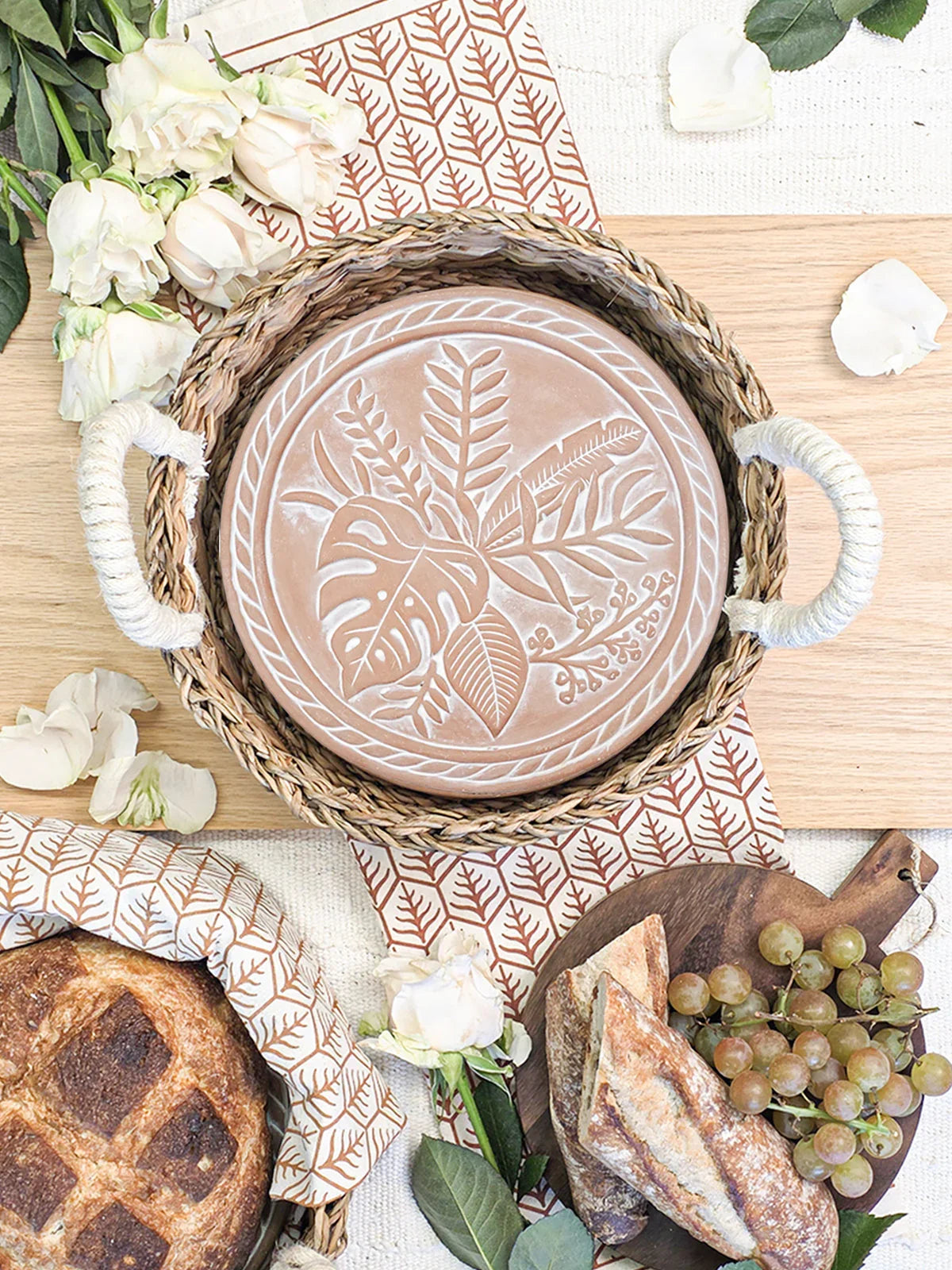 Woven basket with decorative terracotta plate on patterned tea towel, surrounded by bread, grapes, and white roses
