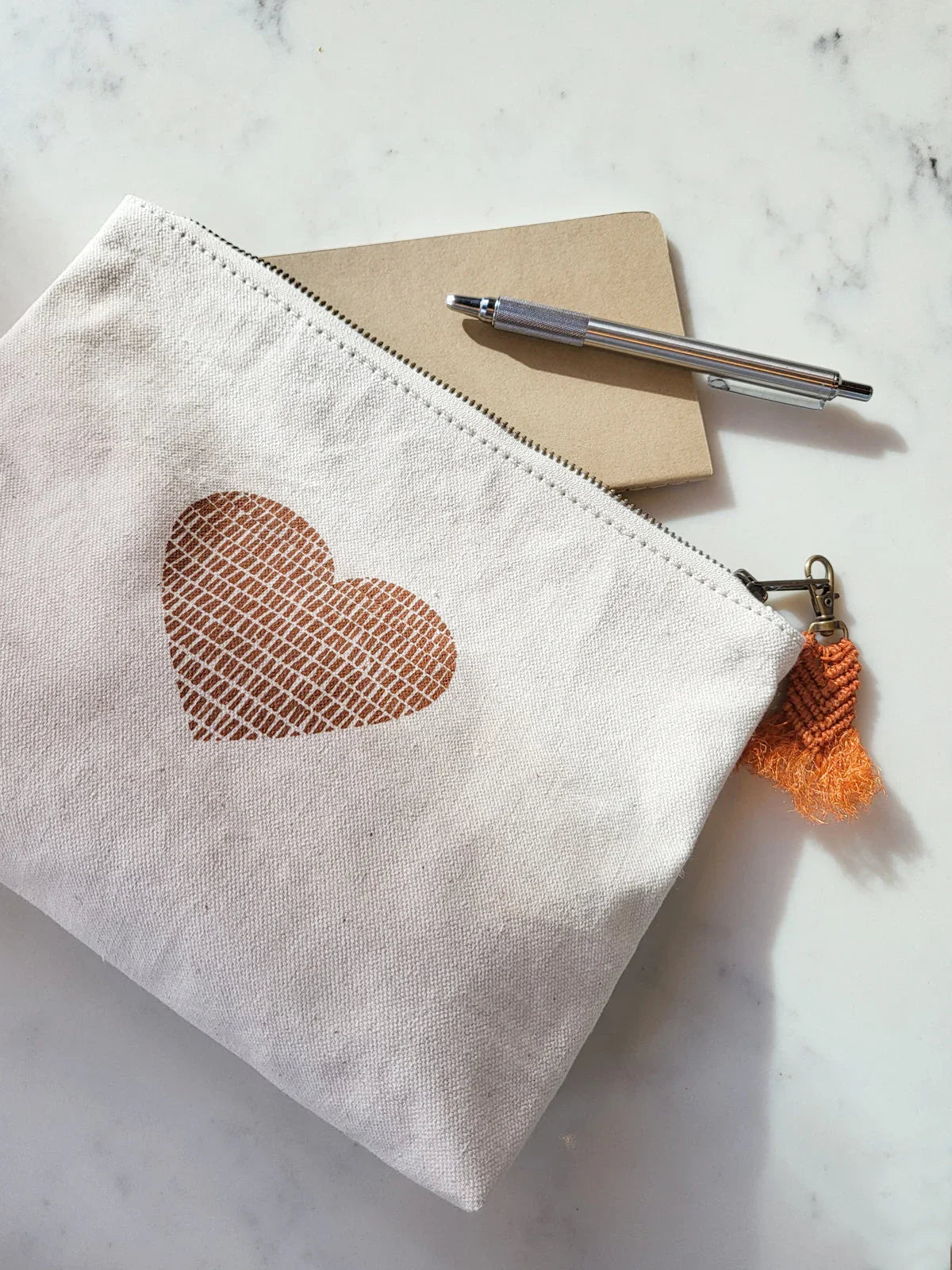White canvas pouch with brown heart print and orange tassel, next to notebook and silver pen on marble surface